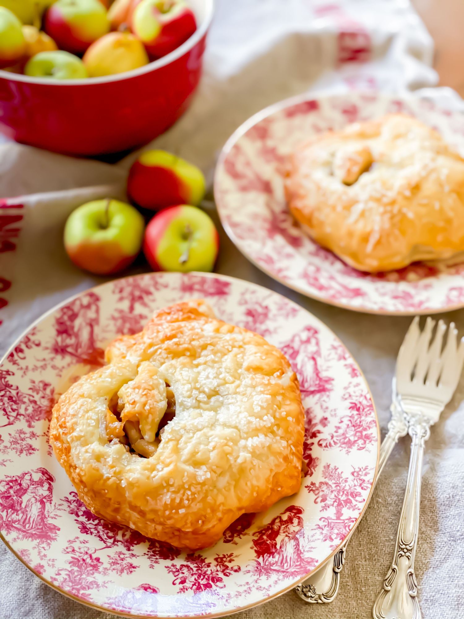 Two golden apple hand pies on red toile plates with fresh apples in a red bowl on a linen tablecloth.