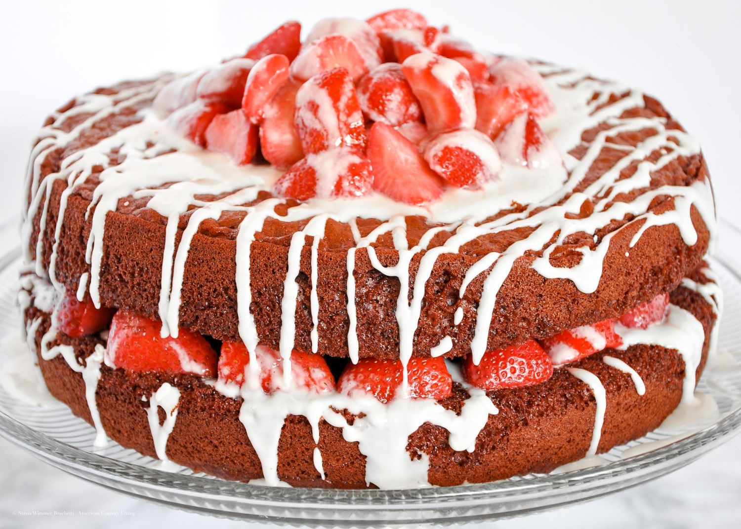 Chocolate layer cake with white icing drizzle and fresh strawberry topping on a glass plate.