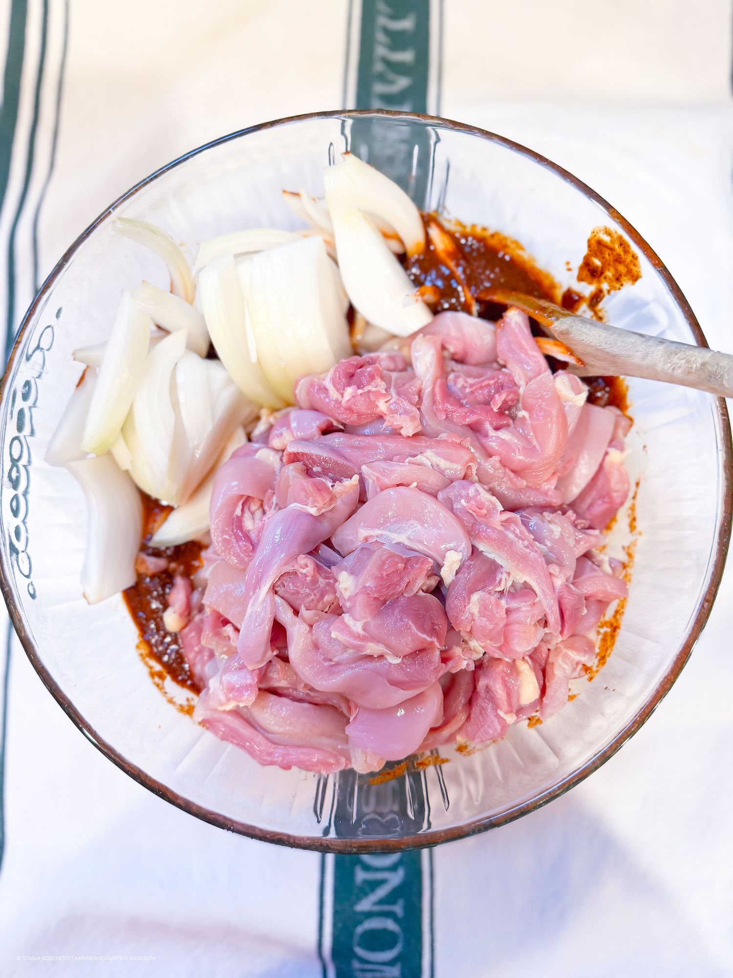 Raw sliced chicken and onions marinating in spicy red sauce in a glass bowl.