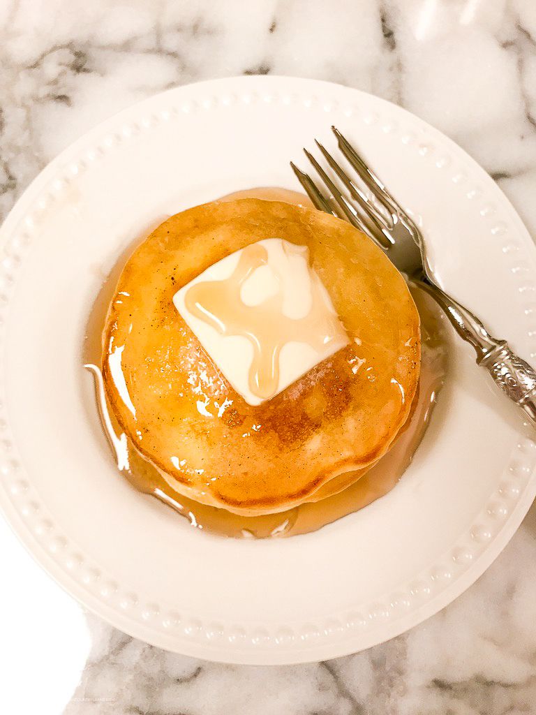 Fluffy pancake topped with melting butter and maple syrup on a white plate with a silver fork.