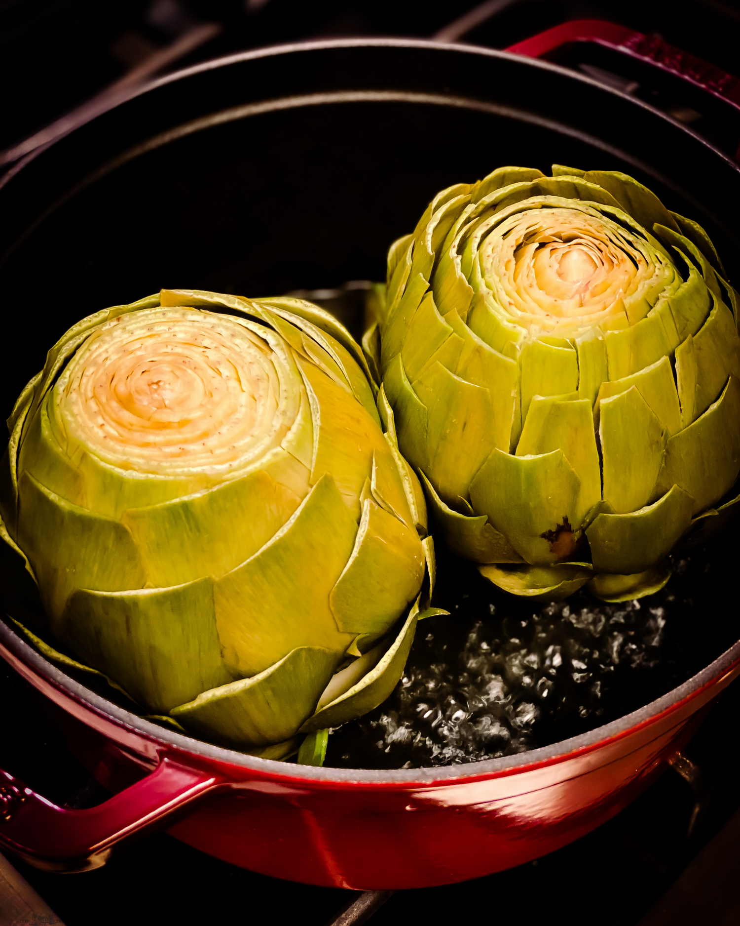 Close-up of a trimmed artichoke head in a red pan with dramatic lighting.