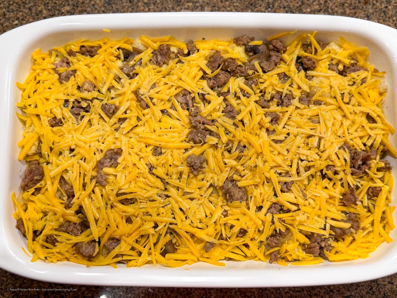 White baking dish filled with seasoned ground beef topped with shredded cheddar cheese, ready for the oven.