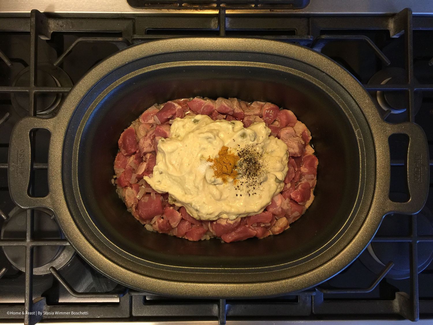 Diced meat with creamy sauce being added to black oval cooking dish.
