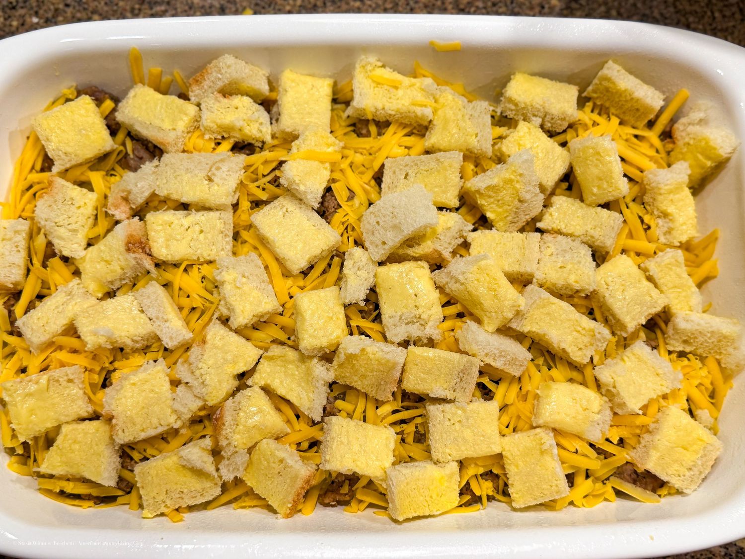 Casserole dish layered with shredded cheddar cheese and cubed bread pieces, ready for baking.
