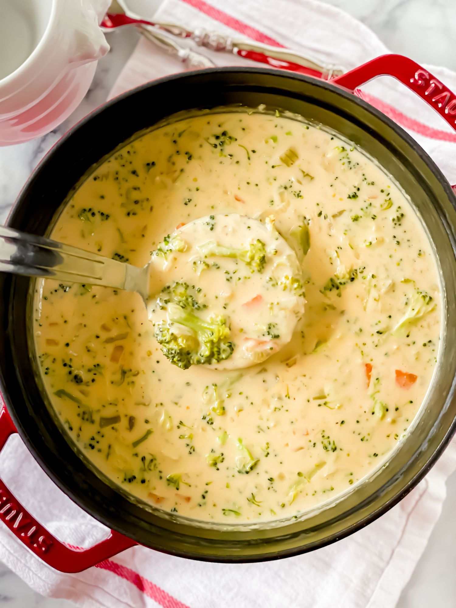 Creamy broccoli cheddar soup in a Staub Dutch oven with a ladle scooping up tender broccoli florets.