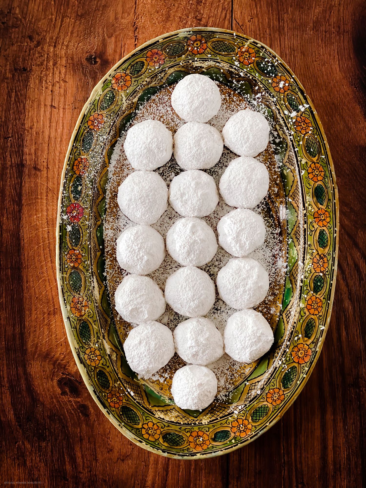 Powdered sugar snowball cookies arranged on a decorative oval floral plate on a wooden surface.