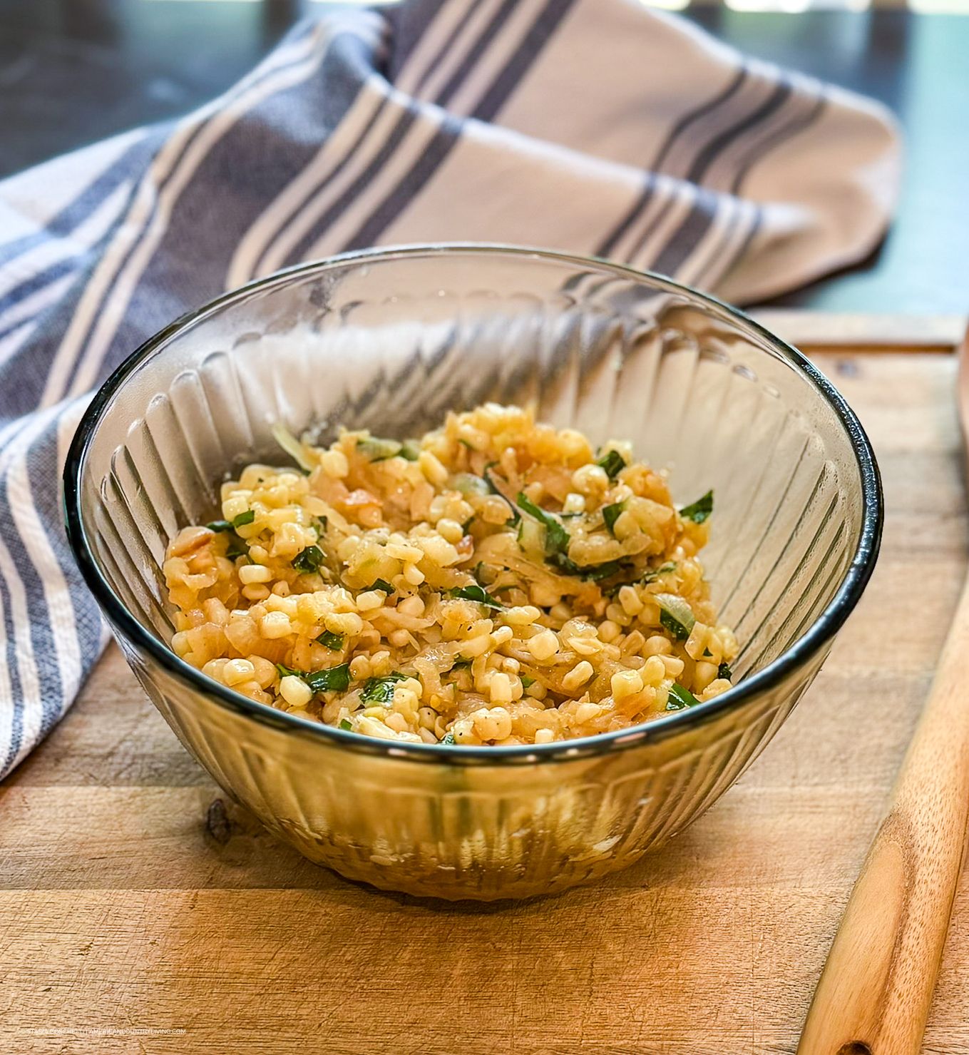 Glass bowl of seasoned bulgur wheat salad with fresh herbs on a wooden cutting board.