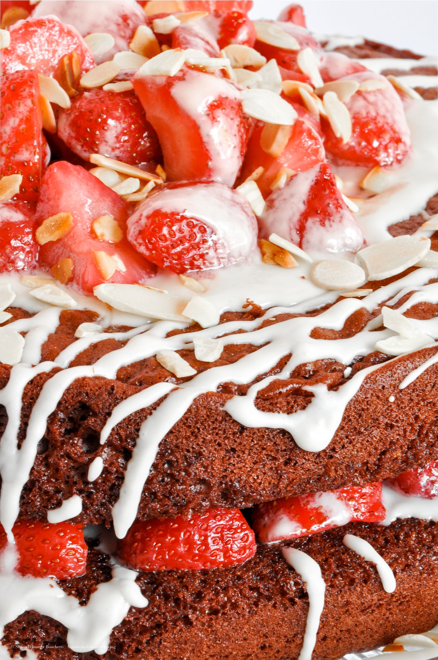 Chocolate layer cake with white glaze, fresh strawberries, and toasted almonds on a glass cake stand.