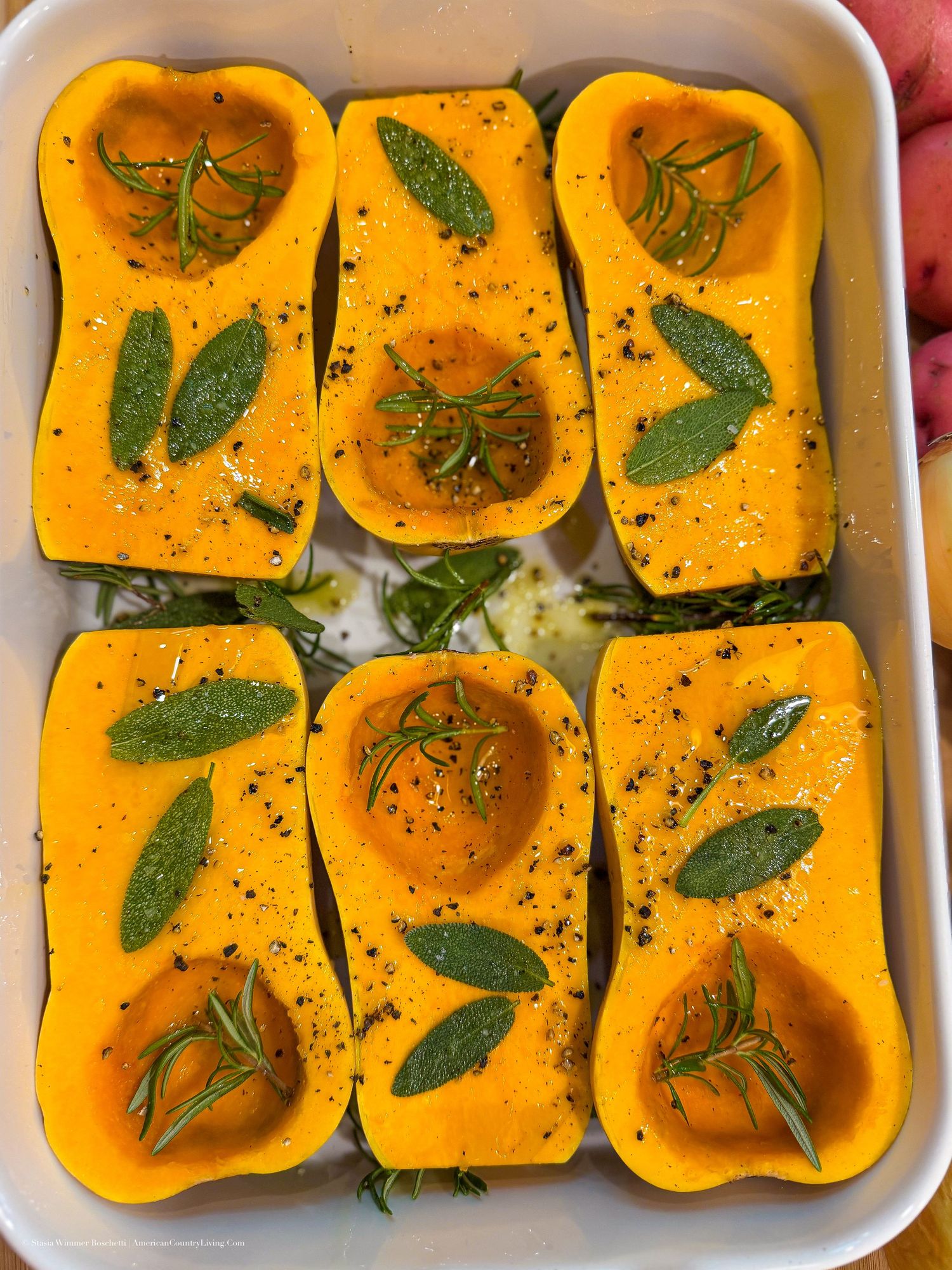 Six halved butternut squash in a white baking dish, topped with fresh sage, rosemary, and black pepper seasoning.