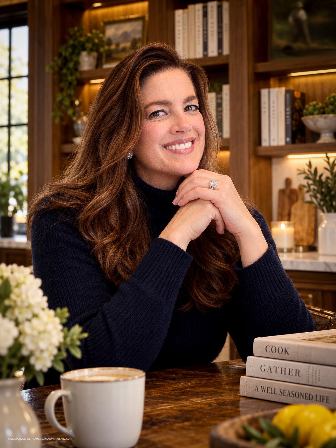 Woman in navy sweater smiling at kitchen counter with coffee, books, and elegant decor in background.