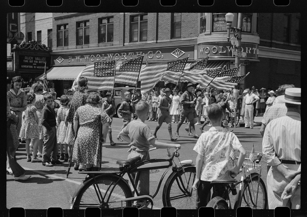 A black and white historical photograph shows a crowded street scene with pedestrians and bicycles in an urban setting.