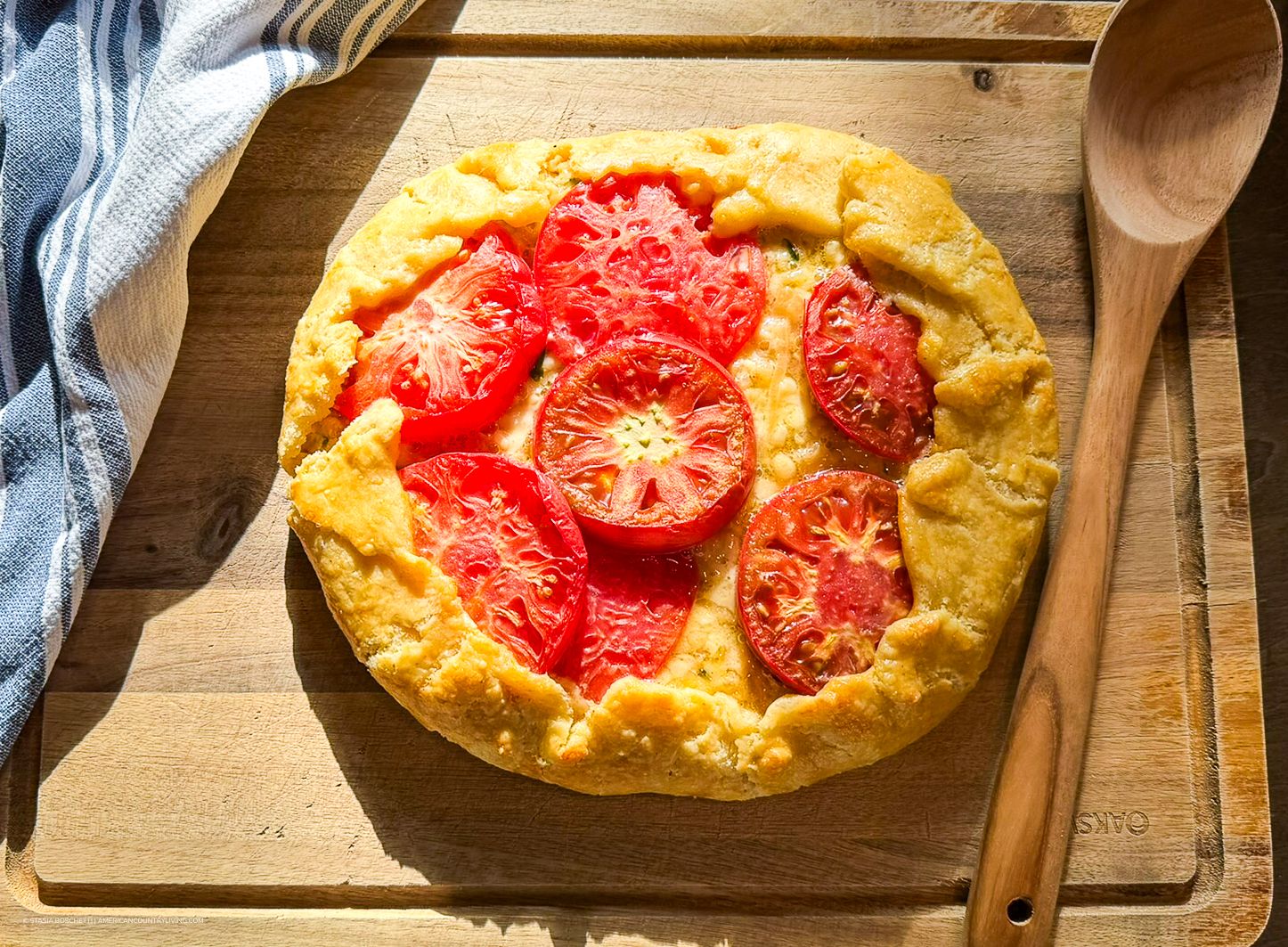 Golden focaccia bread topped with fresh sliced tomatoes on a wooden cutting board with a wooden spoon.