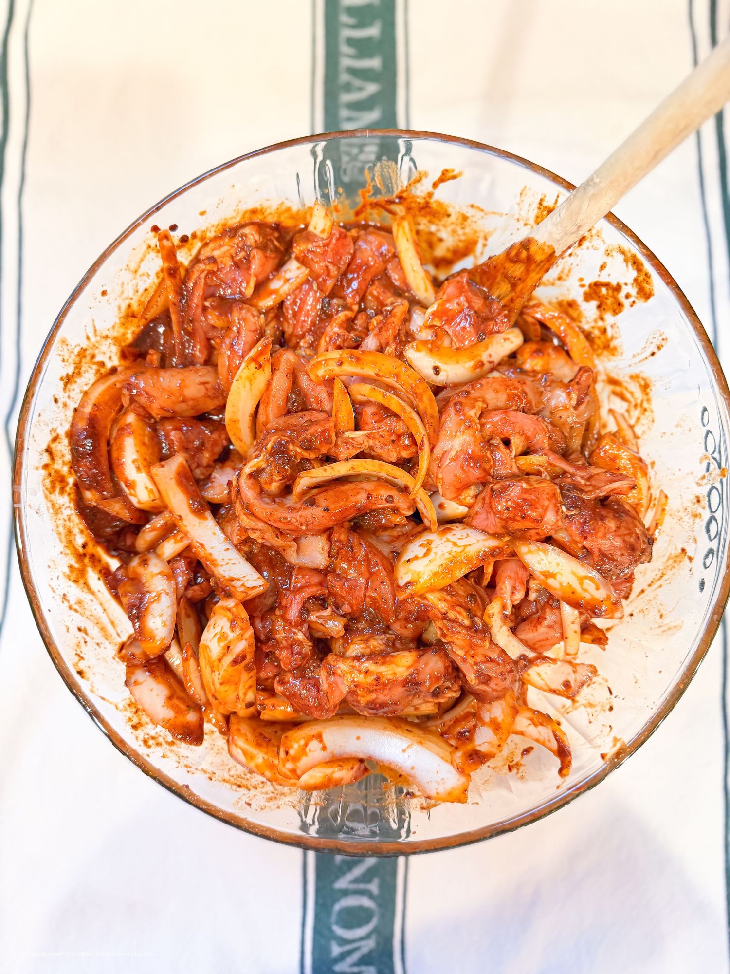 Glass bowl of marinated raw chicken with sliced onions in red spice sauce, mixed with wooden spoon.