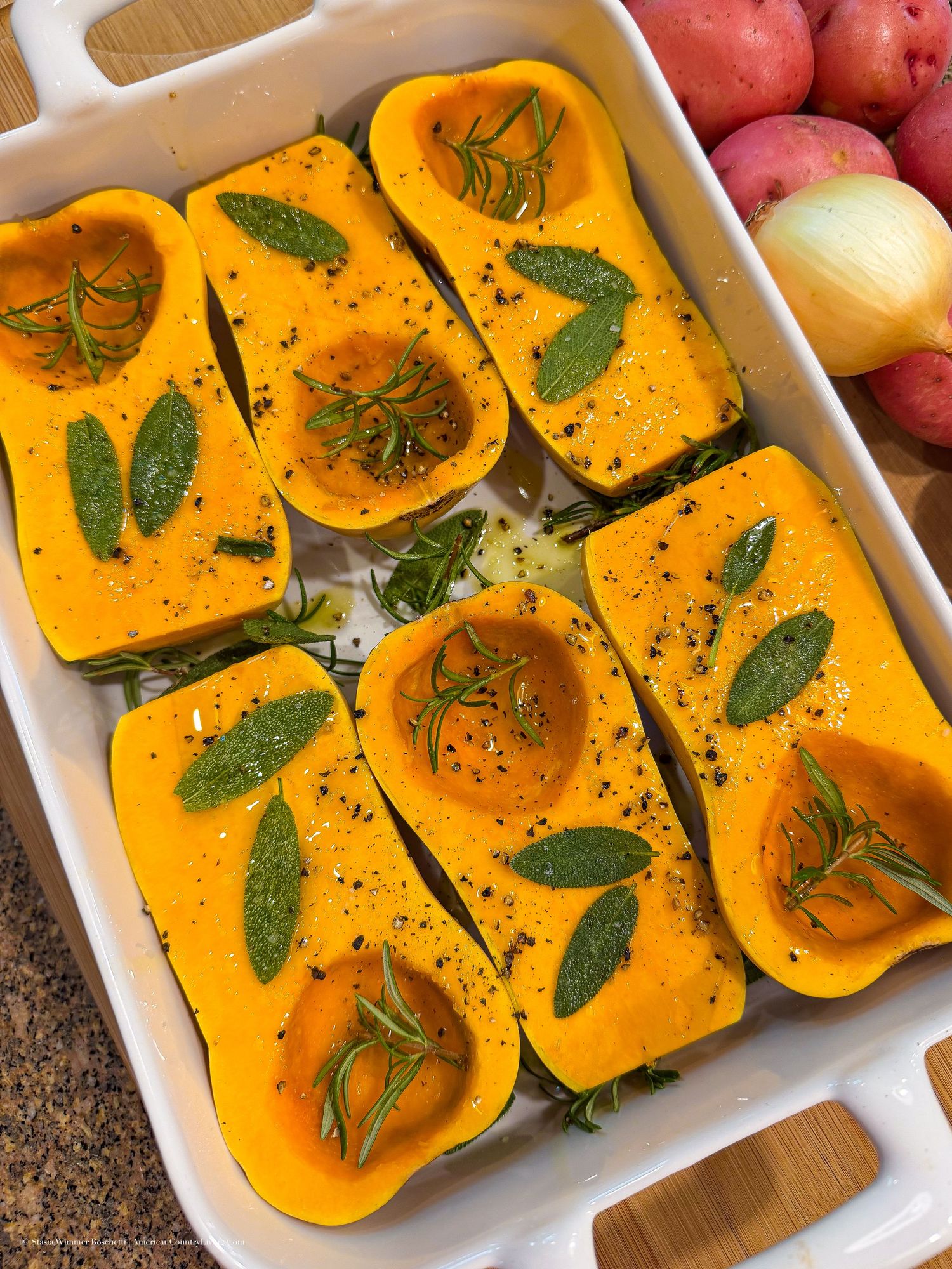 Halved butternut squash seasoned with fresh sage and rosemary in a white baking dish, ready to roast.