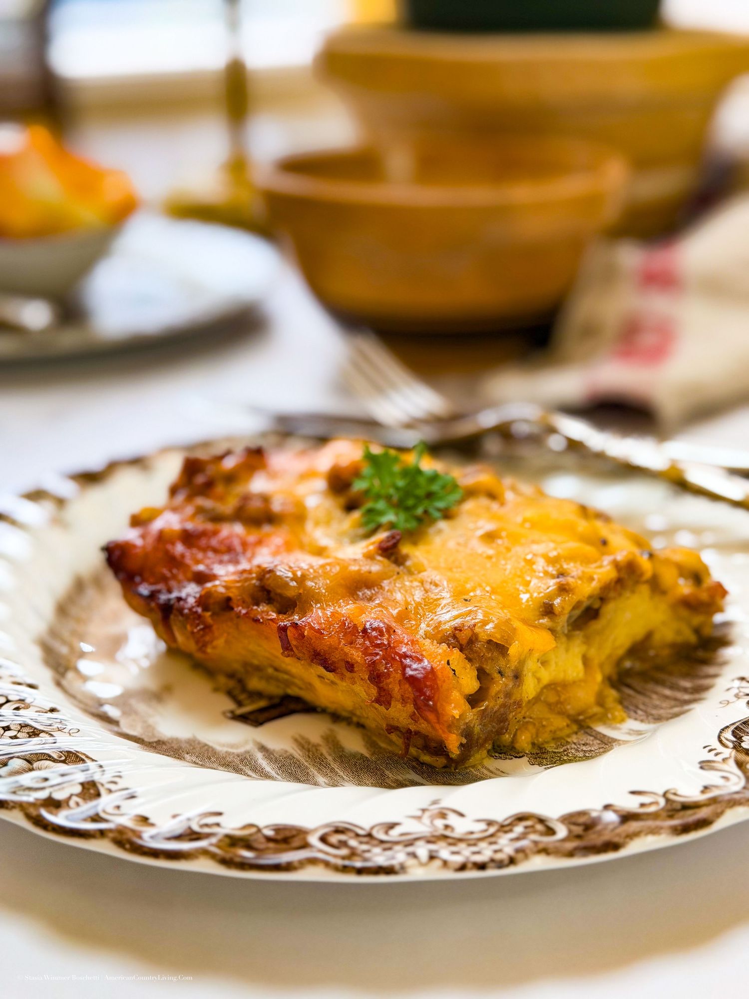 A golden-brown slice of cheesy lasagna served on an ornate white plate with a fork in the background.