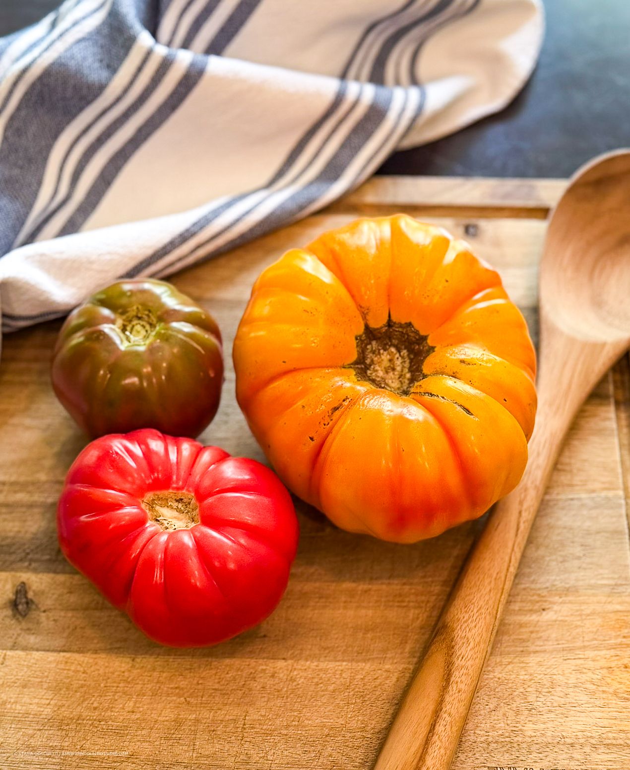 Three colorful heirloom tomatoes on a wooden cutting board with a wooden spoon and striped cloth napkin.