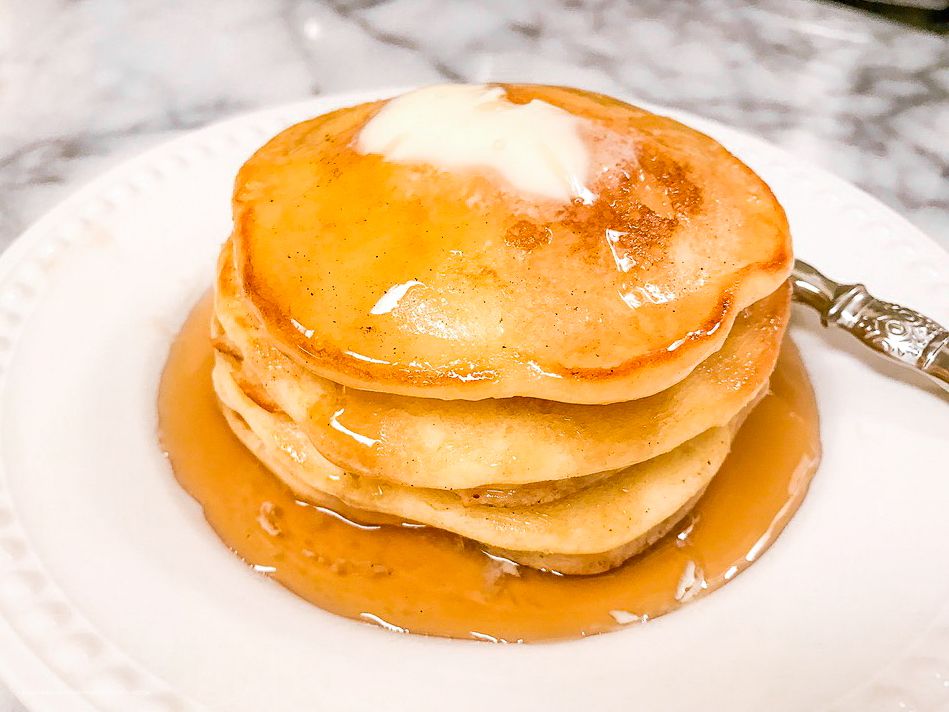 Stack of fluffy golden pancakes topped with butter and maple syrup on a white plate.