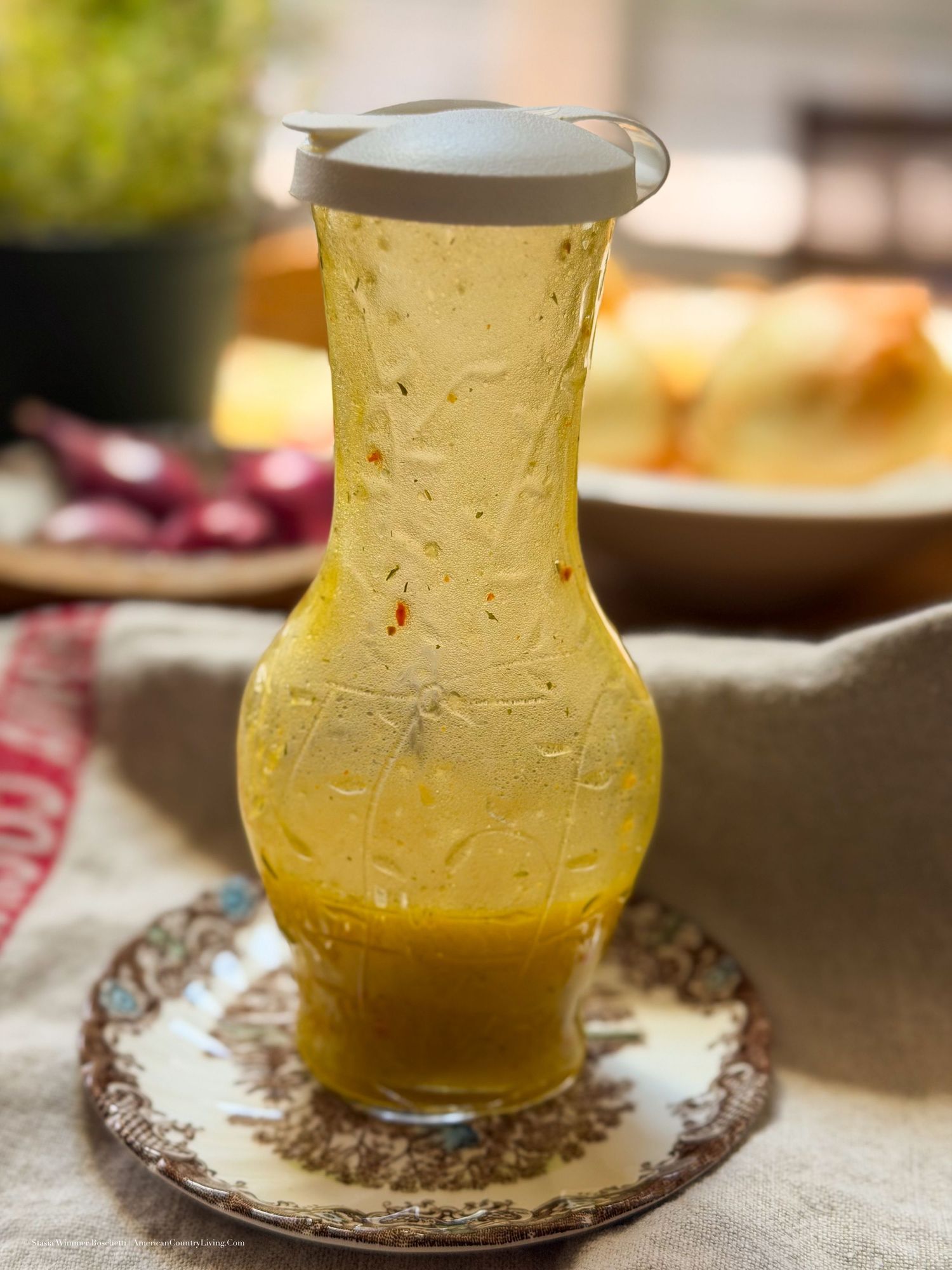 A decorative glass pitcher filled with golden yellow salad dressing sits on an ornate saucer on a linen tablecloth.