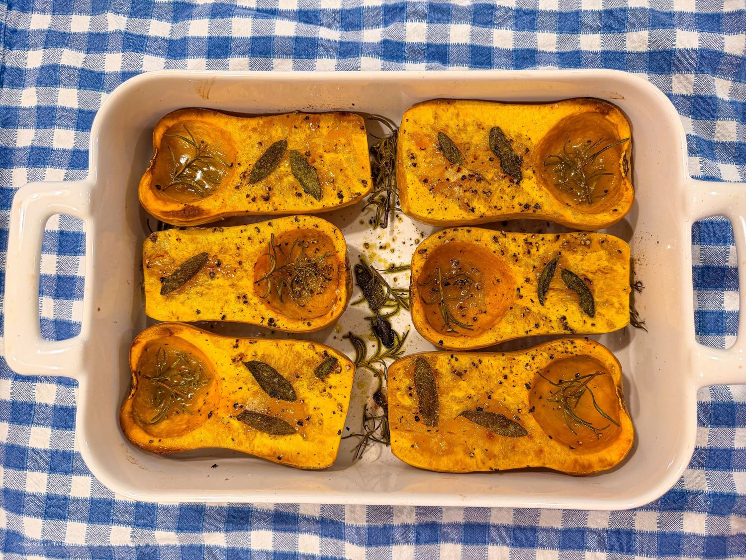 Roasted butternut squash halves with herbs and olive oil in a white baking dish on a blue checkered cloth.