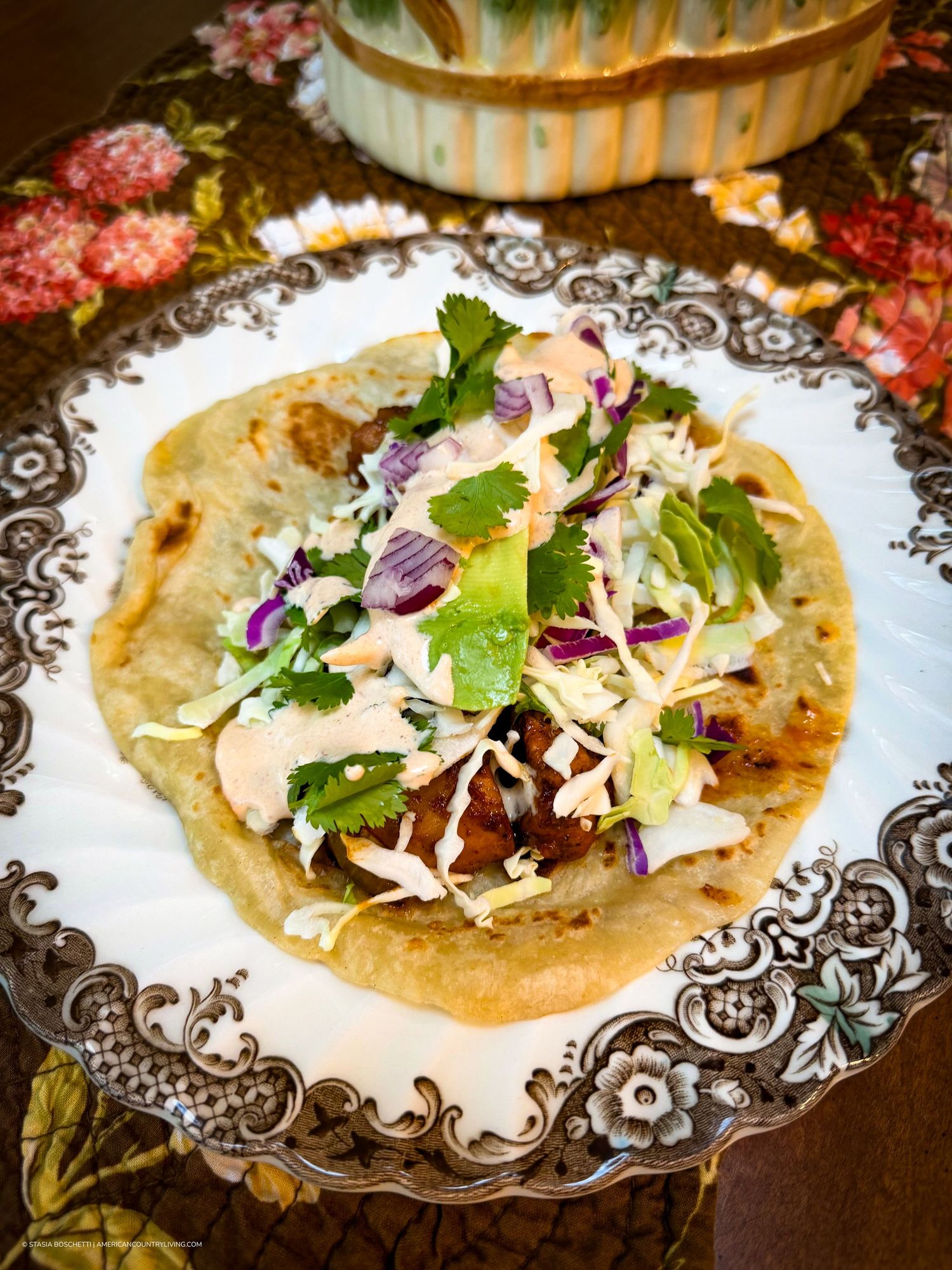 A gourmet fish taco on a flour tortilla topped with coleslaw, cilantro, red cabbage, and chipotle sauce on a decorative plate.