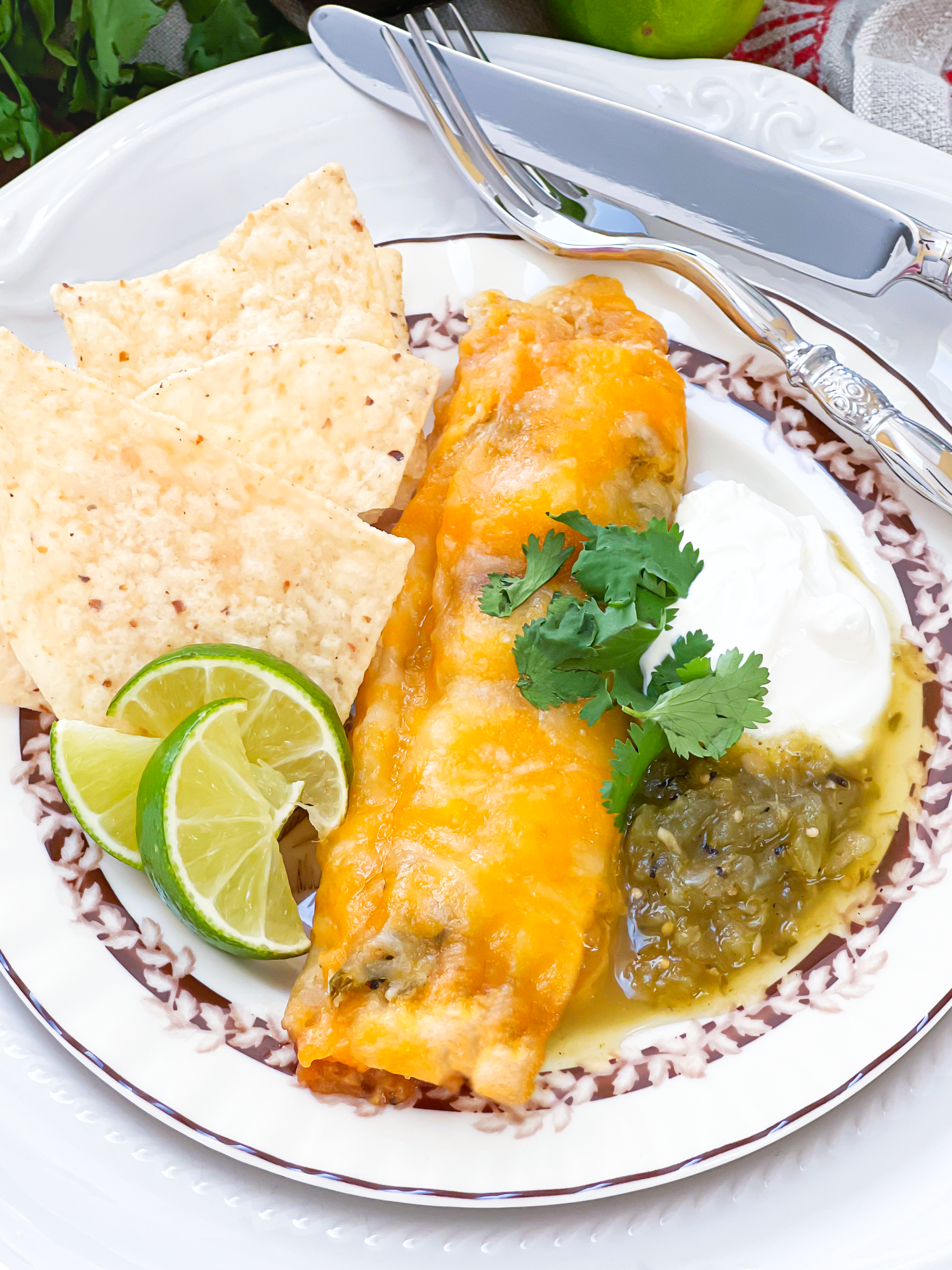 Cheesy enchiladas served with lime wedges, salsa verde, and tortilla chips on a decorative plate.