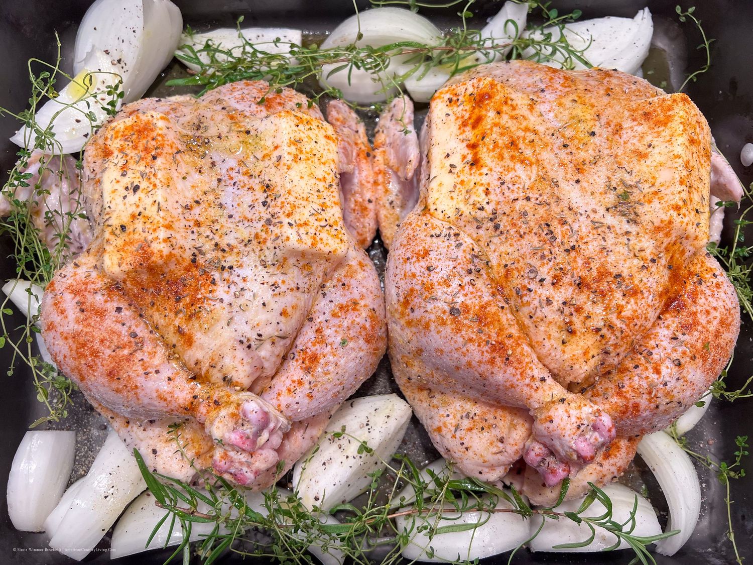 Seasoned chicken thighs nestled among onions and fresh herbs in a dark roasting dish.