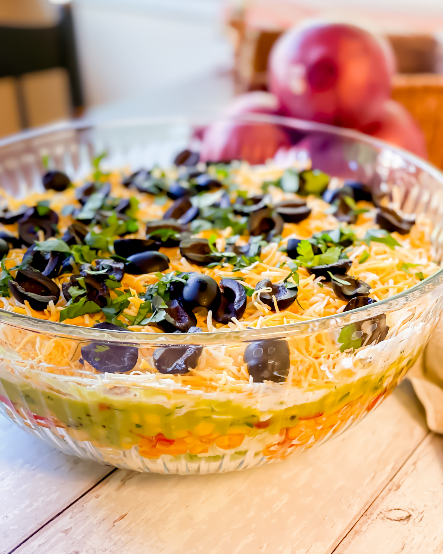 A layered dip in a glass dish topped with shredded cheese, black olives, and fresh herbs.