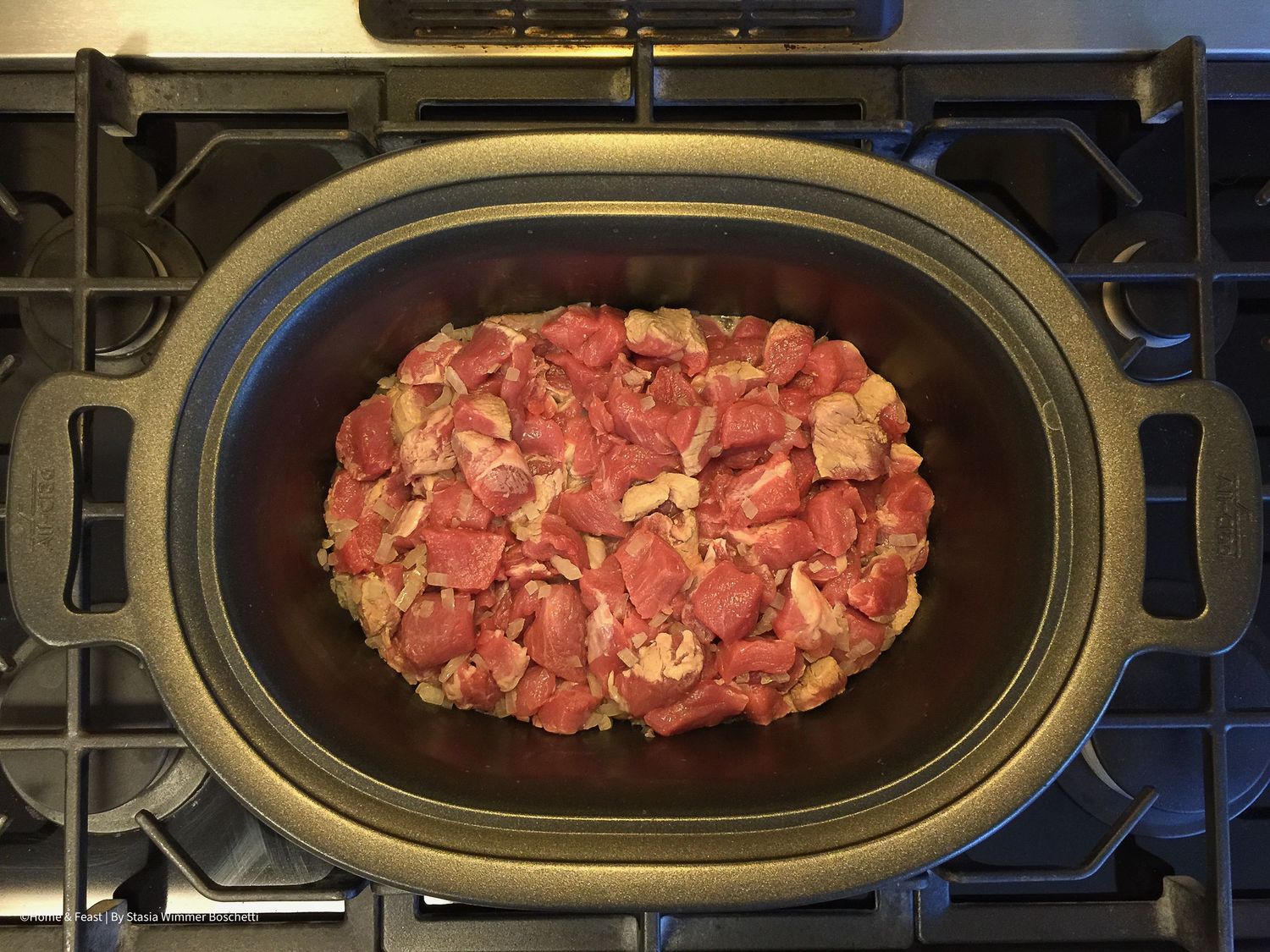 Raw diced meat arranged in an oval-shaped black cooking dish on a stovetop.