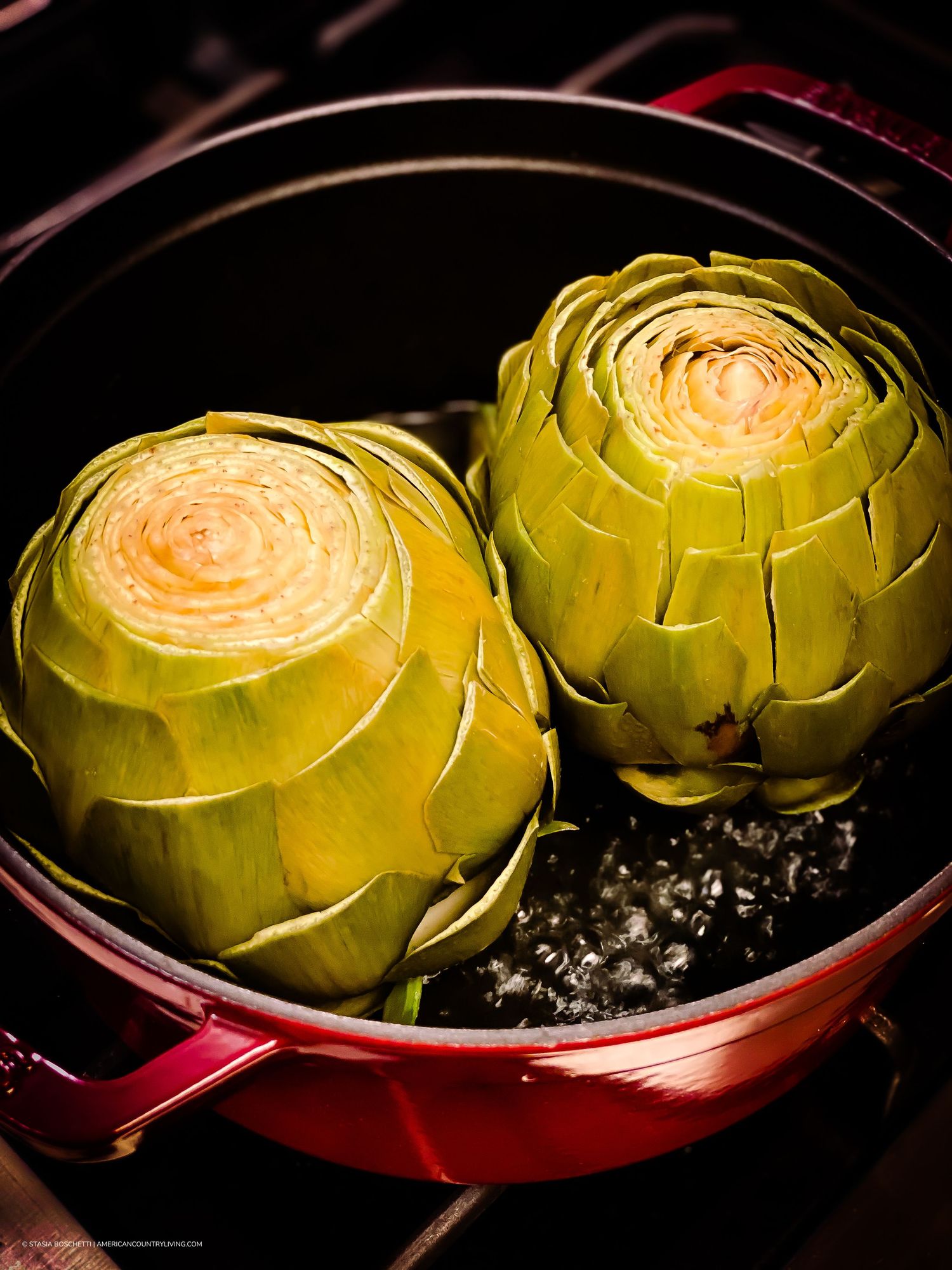 Two fresh artichokes steaming in a red Dutch oven pot on a stovetop burner.
