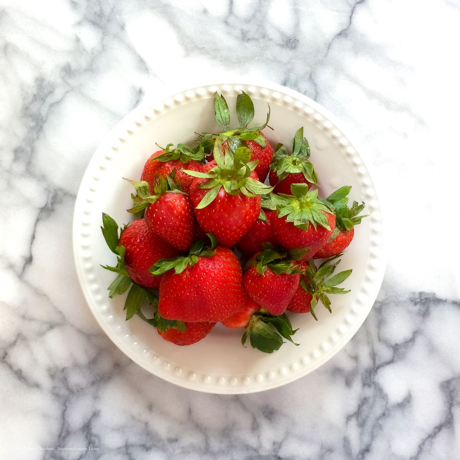 Fresh red strawberries with green stems piled in a white ceramic bowl on a marble surface.