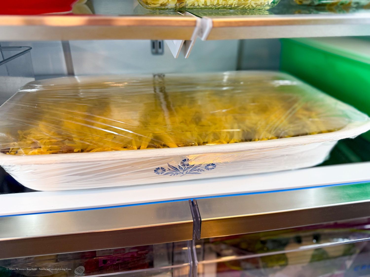 Corelle dish covered with plastic wrap storing yellow pasta in a refrigerator on glass shelves.