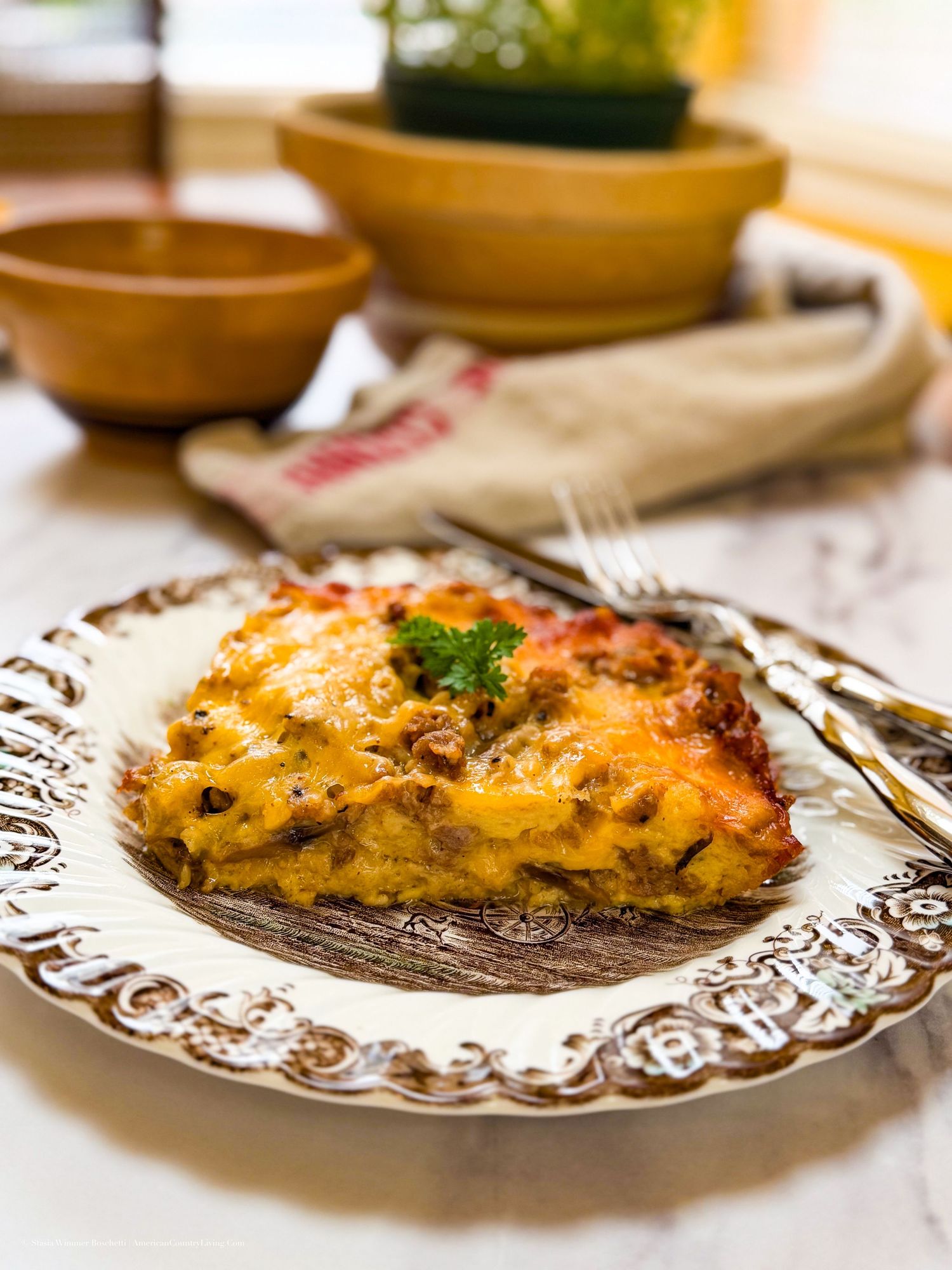 A golden slice of cheesy lasagna on an ornate white plate, garnished with parsley, with rustic bowls in the background.