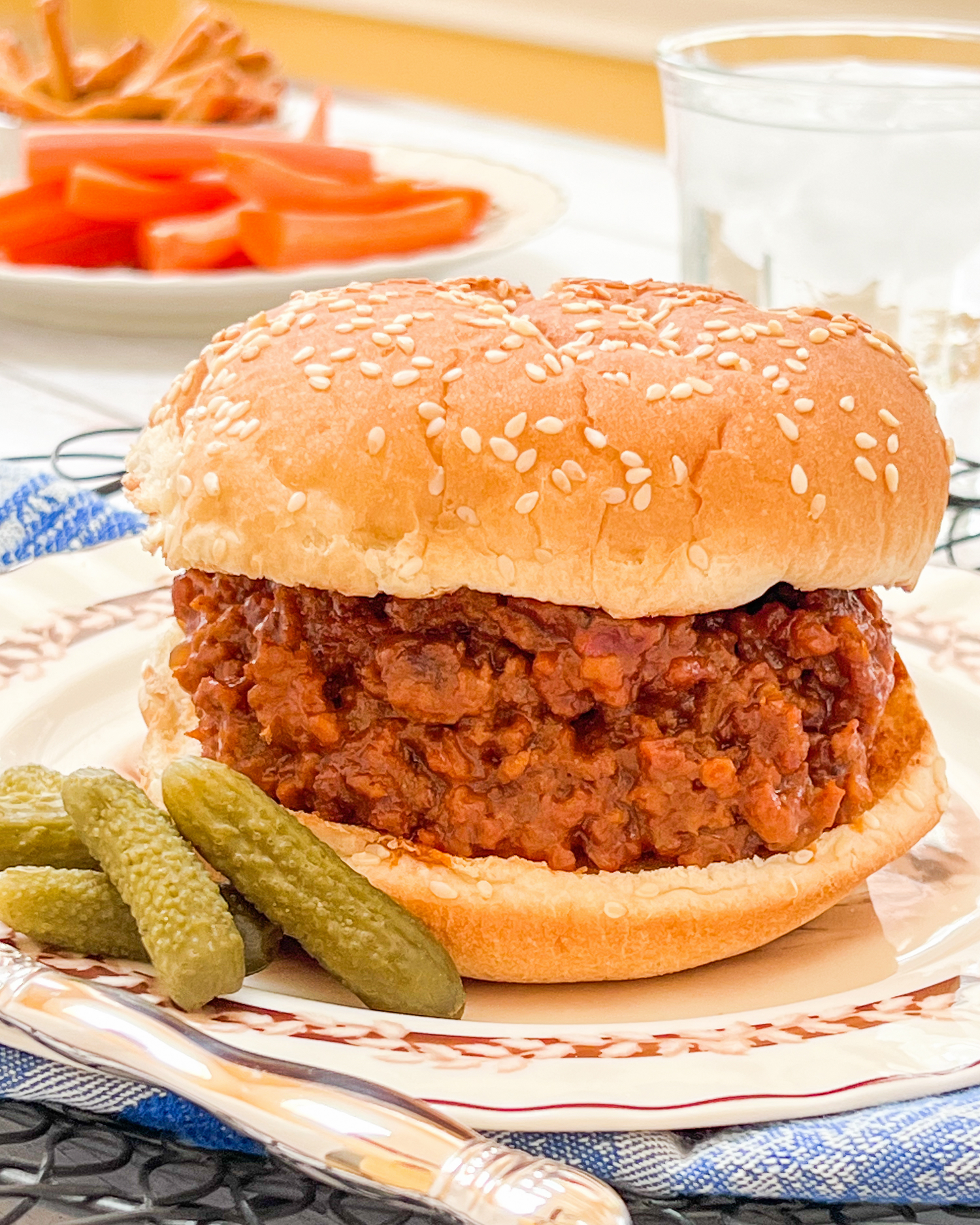 Juicy sloppy joe sandwich on a toasted bun with sesame seeds, served with dill pickles and fresh carrots.
