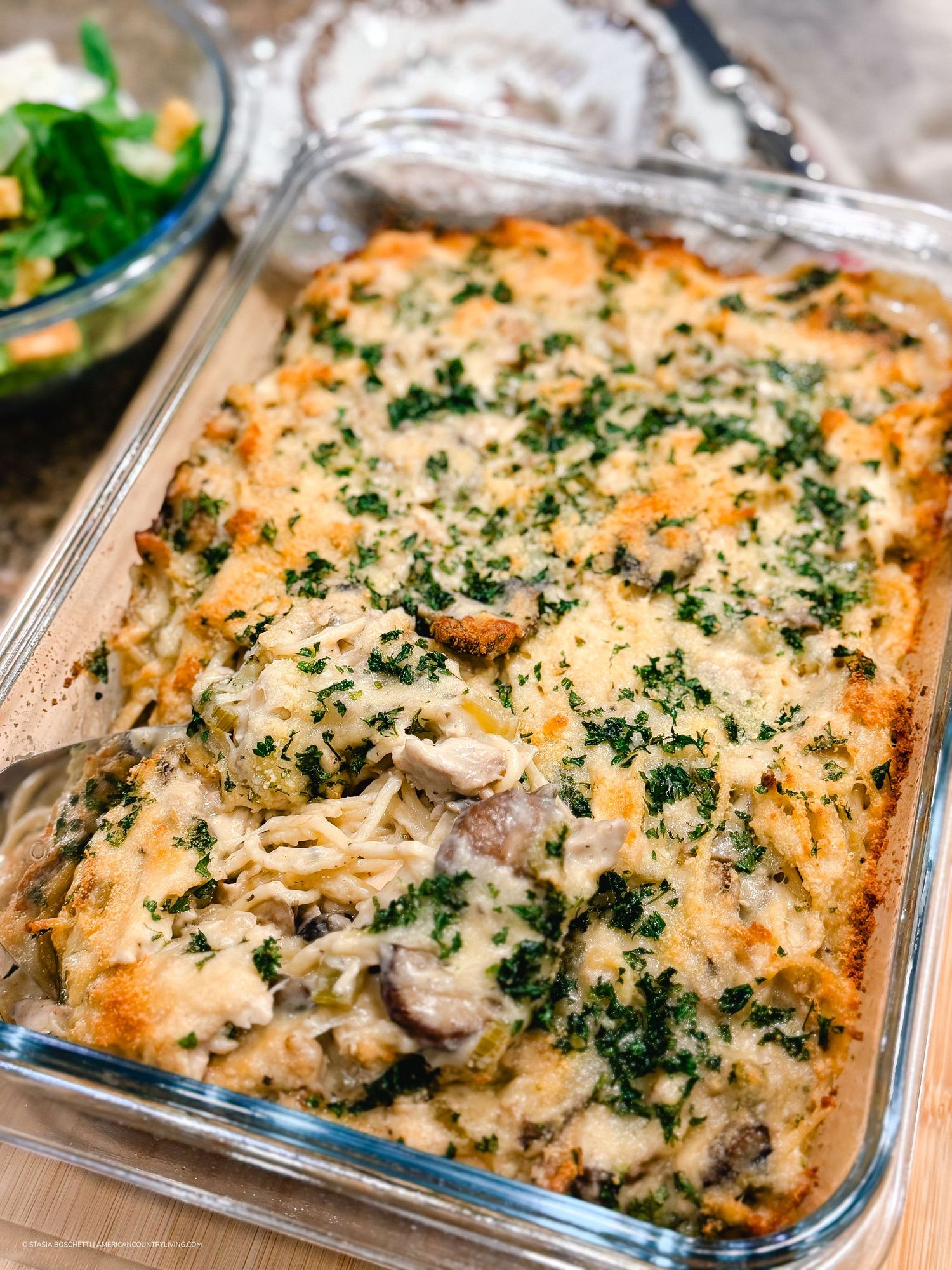 Golden cheesy chicken and mushroom casserole topped with herbs, baked in a glass dish, served alongside a fresh green salad.
