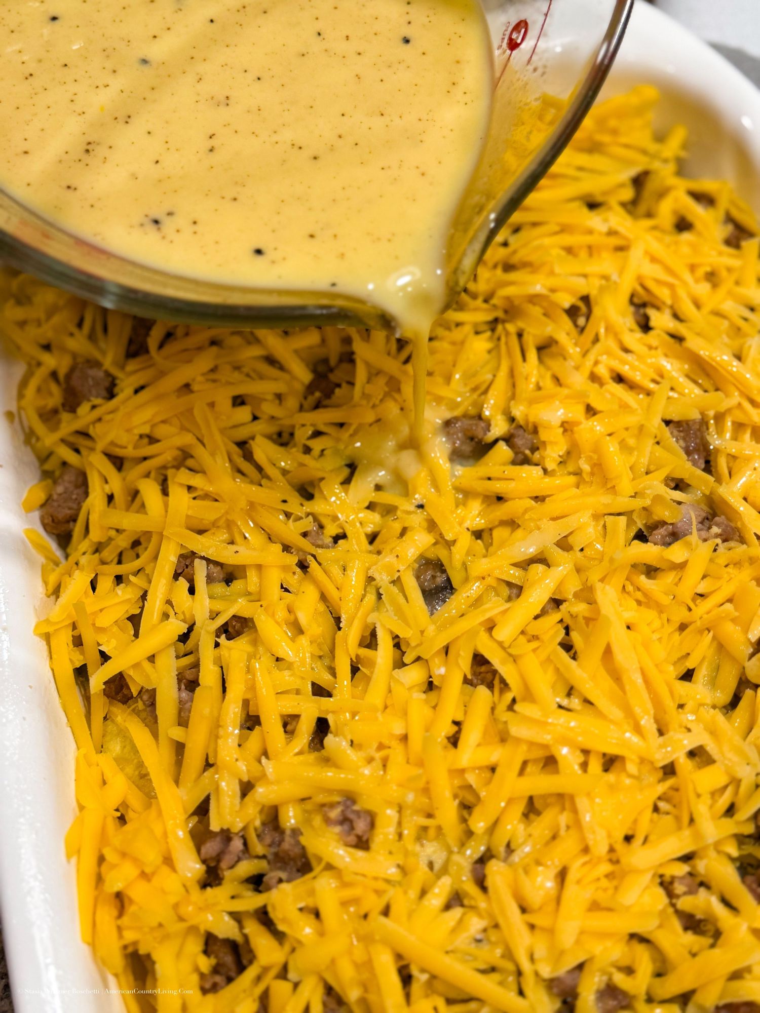 Cheesy casserole dish with shredded cheddar and ground beef, topped with creamy sauce being poured from a glass measuring cup.