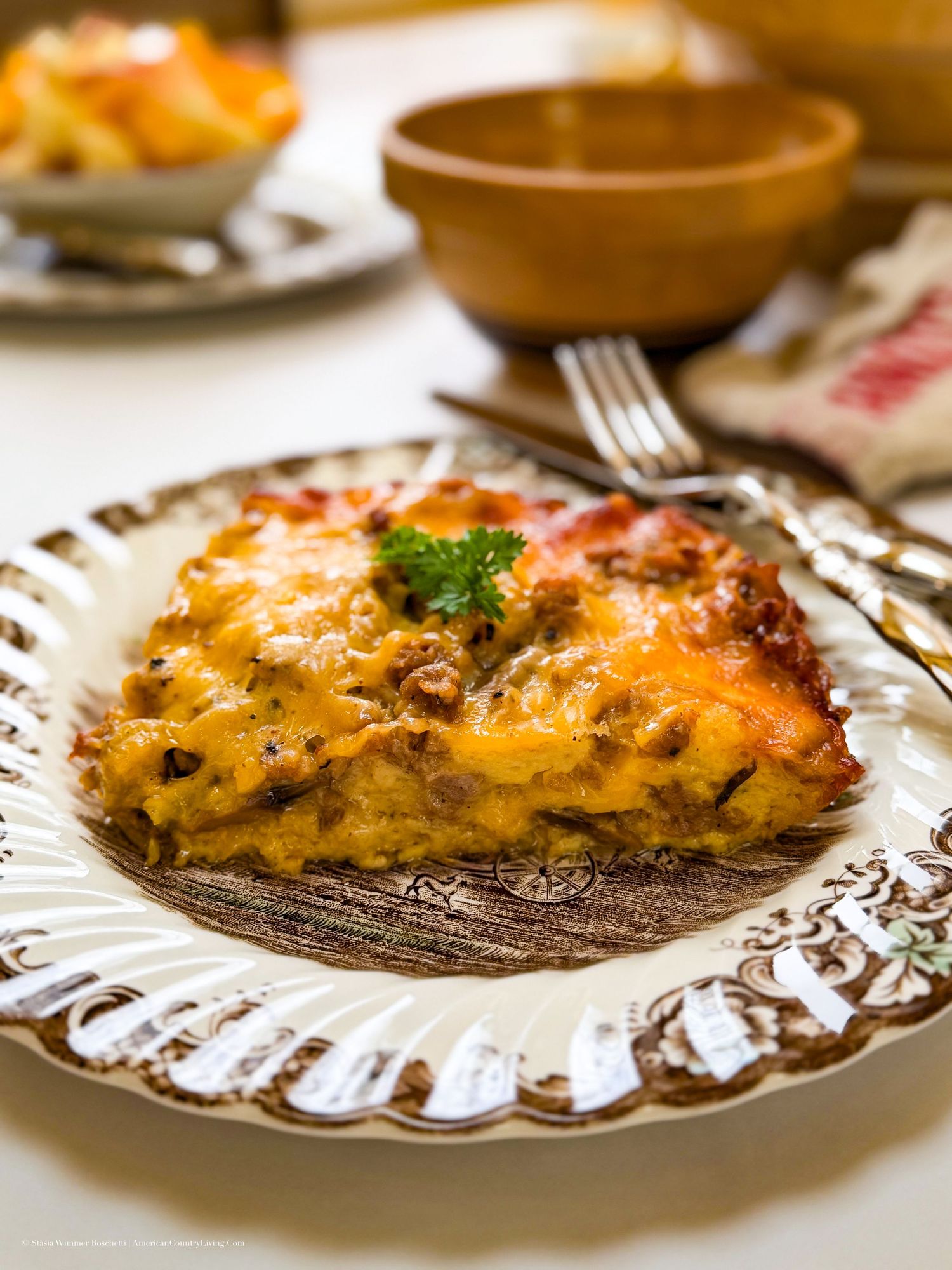 A golden-brown cheesy pasta bake slice on a decorative plate, garnished with fresh parsley, served on a white tablecloth.