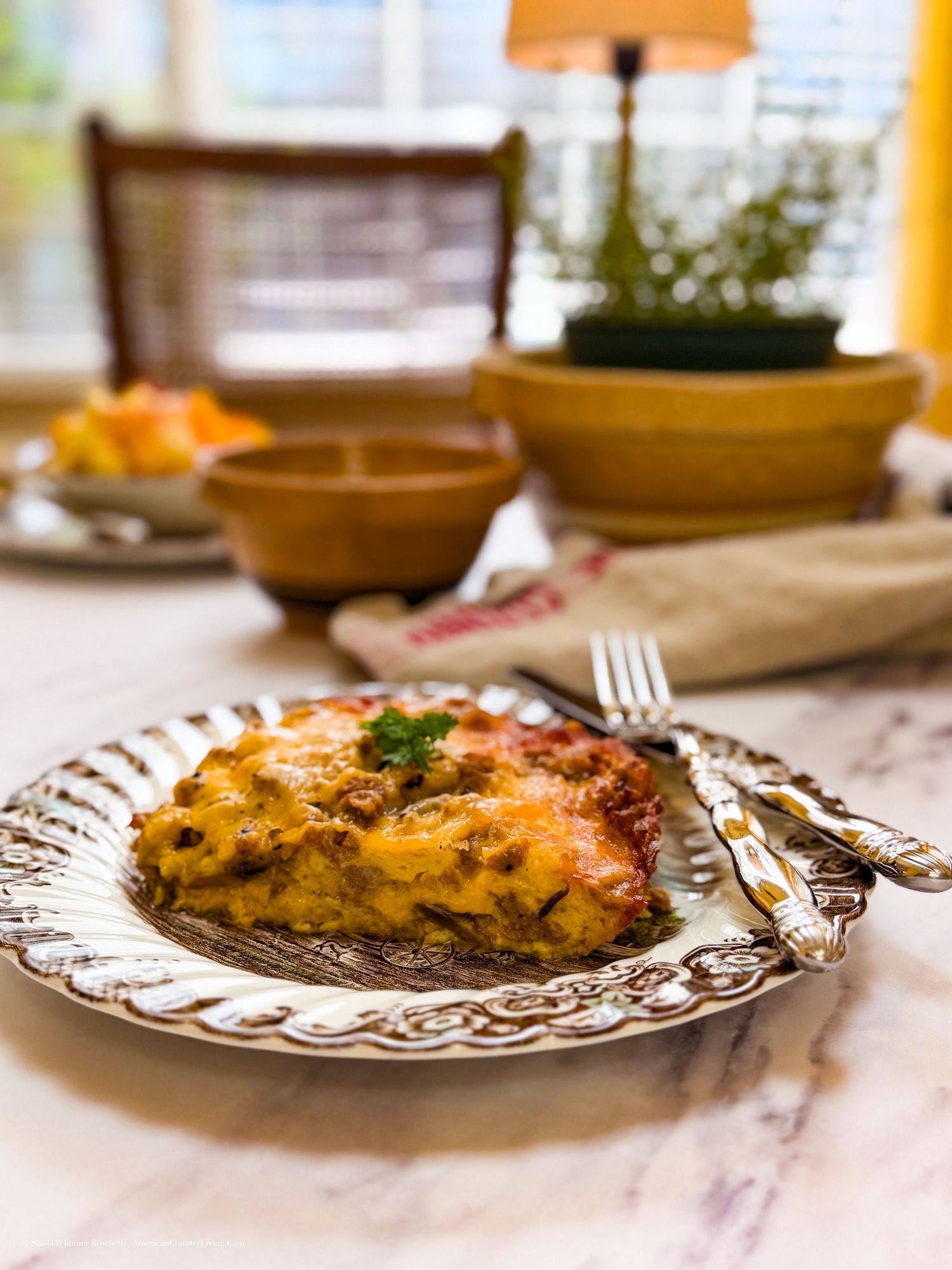 A slice of cheesy lasagna on a decorative silver plate with a fork, served on a marble table in a cozy dining setting.
