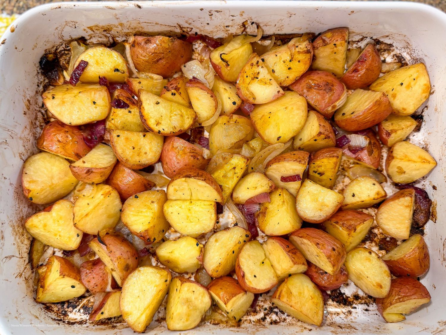 Golden roasted potatoes with red onions, herbs, and spices baked in a white casserole dish.