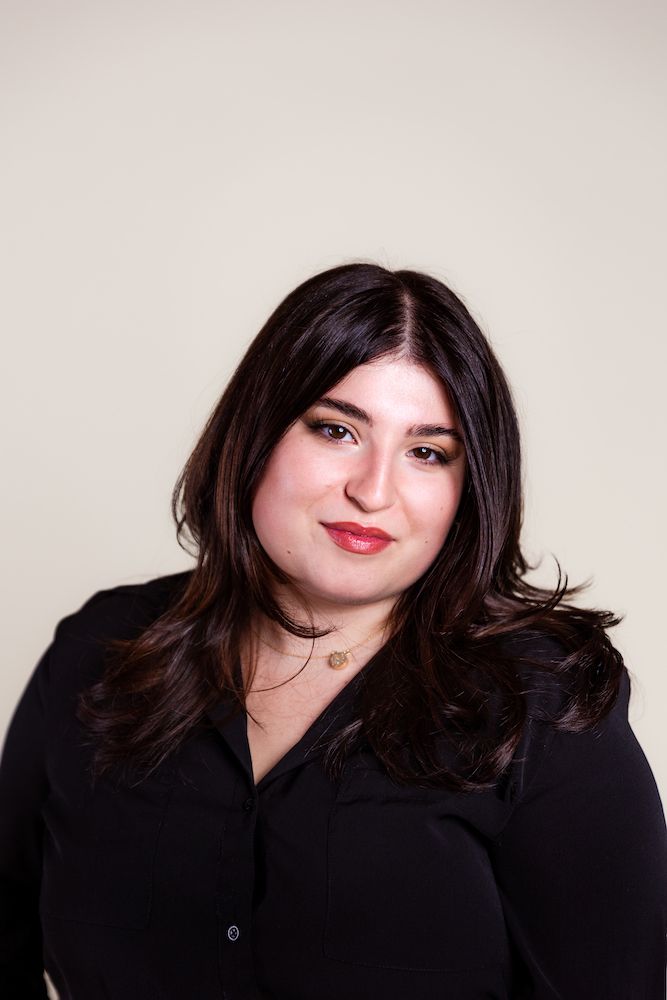 Young brunette woman in black blouse with red lips smiling softly against a light neutral background.