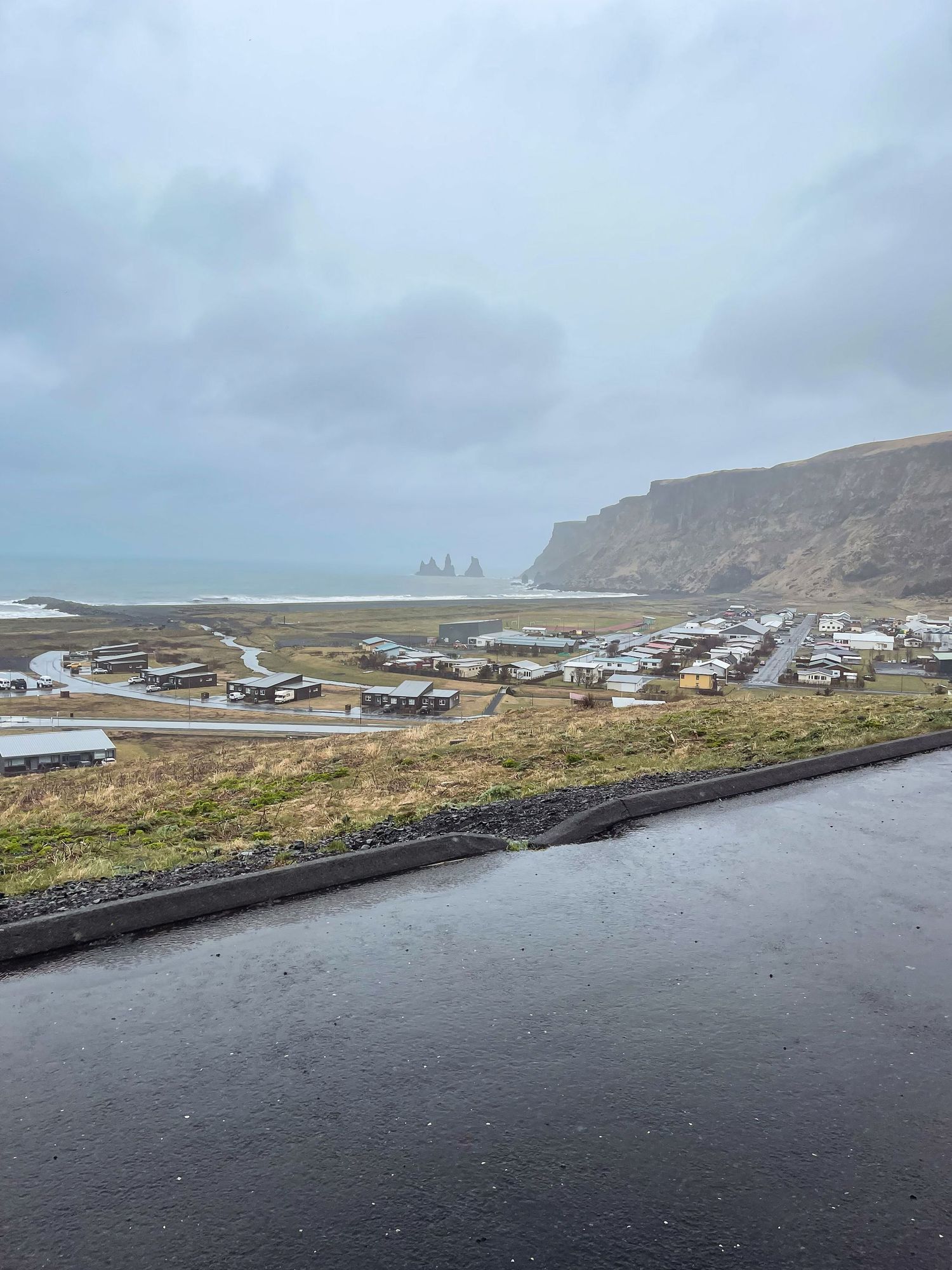 Coastal village nestled beneath cliffs with famous Reynisdrangar sea stacks visible in the distance.