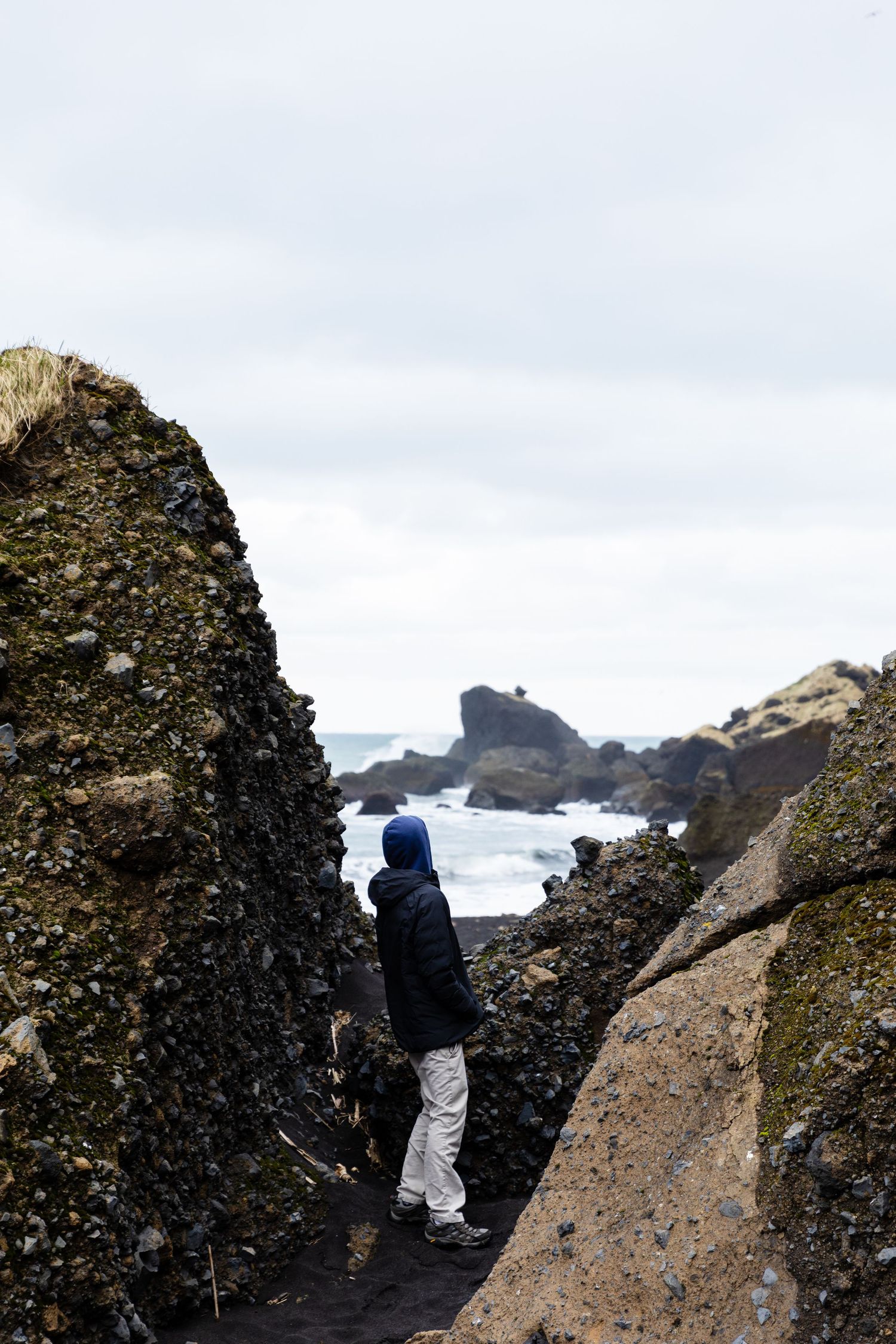 Figure in blue hoodie stands between rocky cliffs overlooking a dramatic coastal landscape in Iceland.