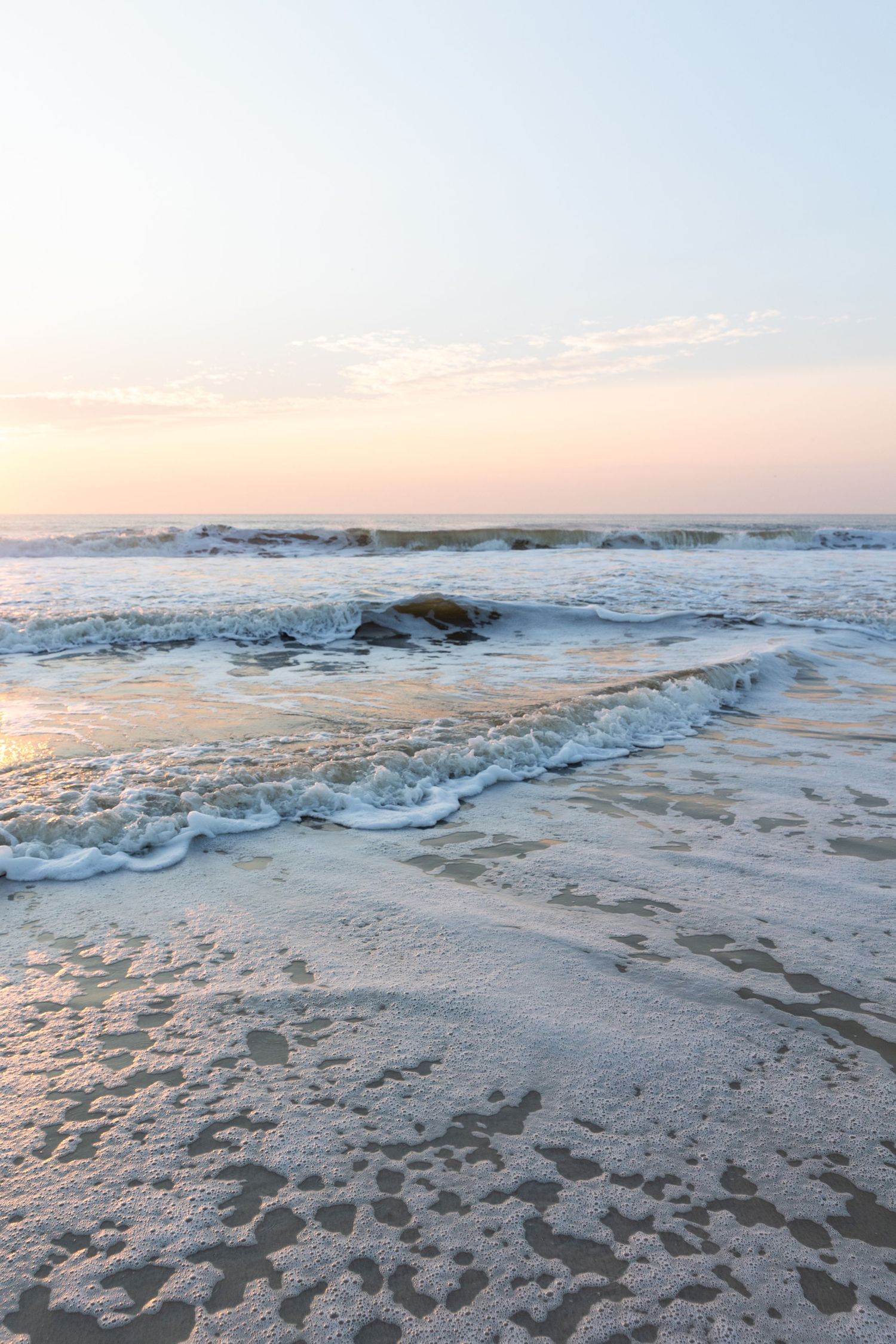 Ocean waves crash onto sandy beach during sunset creating a serene coastal scene with soft pastel colors.