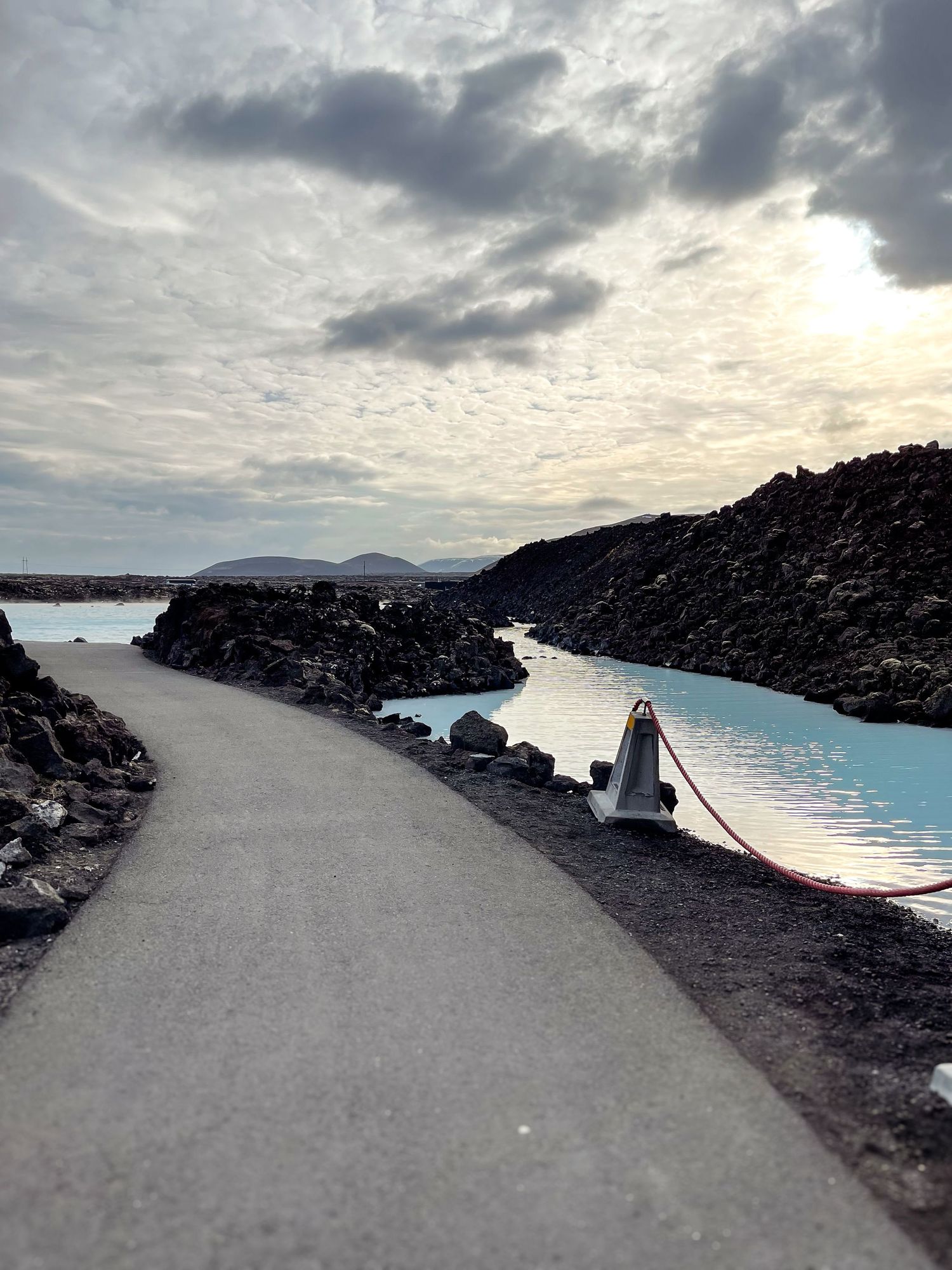 Path winding between black rocks and bright blue thermal waters under cloudy sky.