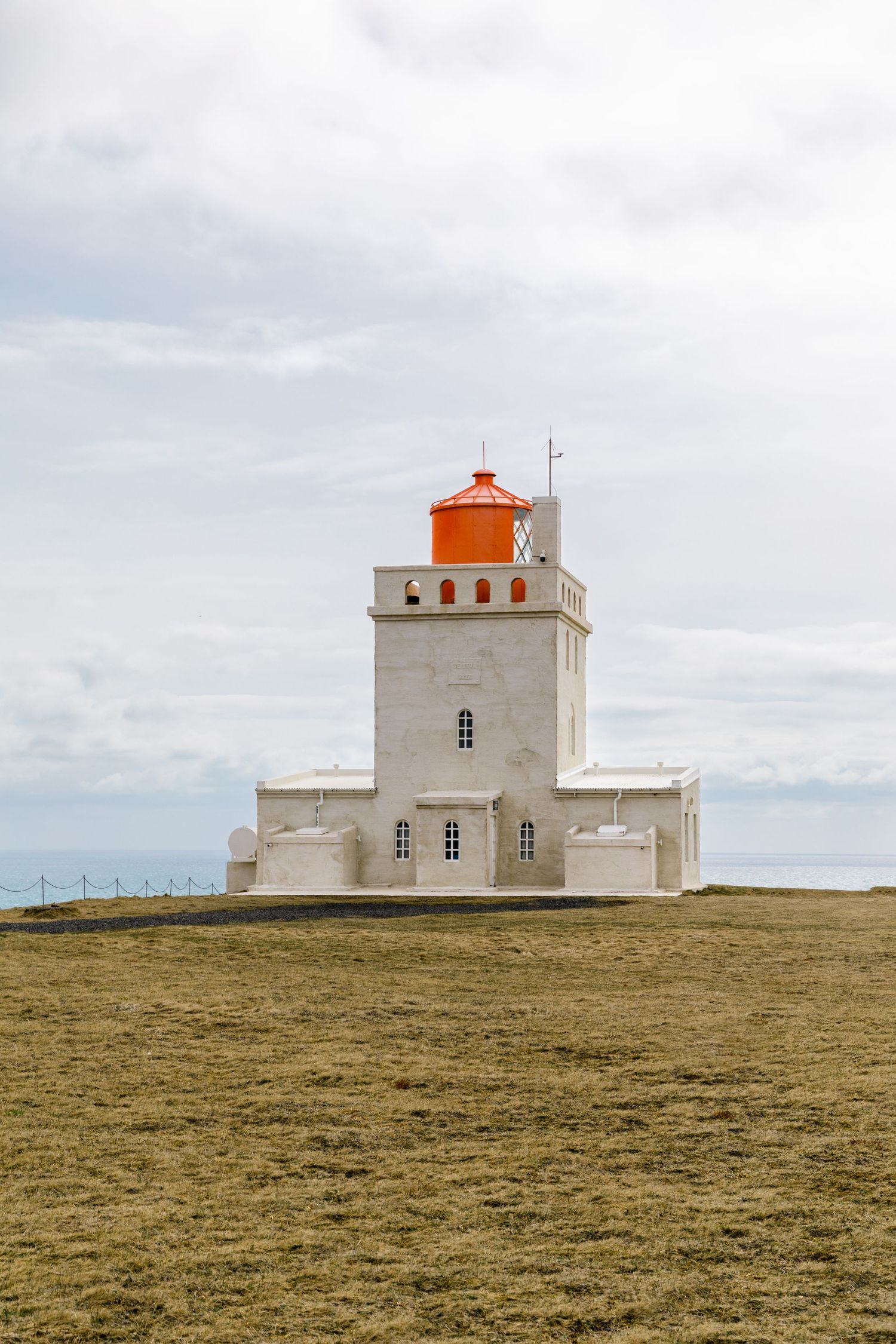 White lighthouse with orange top stands solitary on grassy coastal cliff in Iceland.