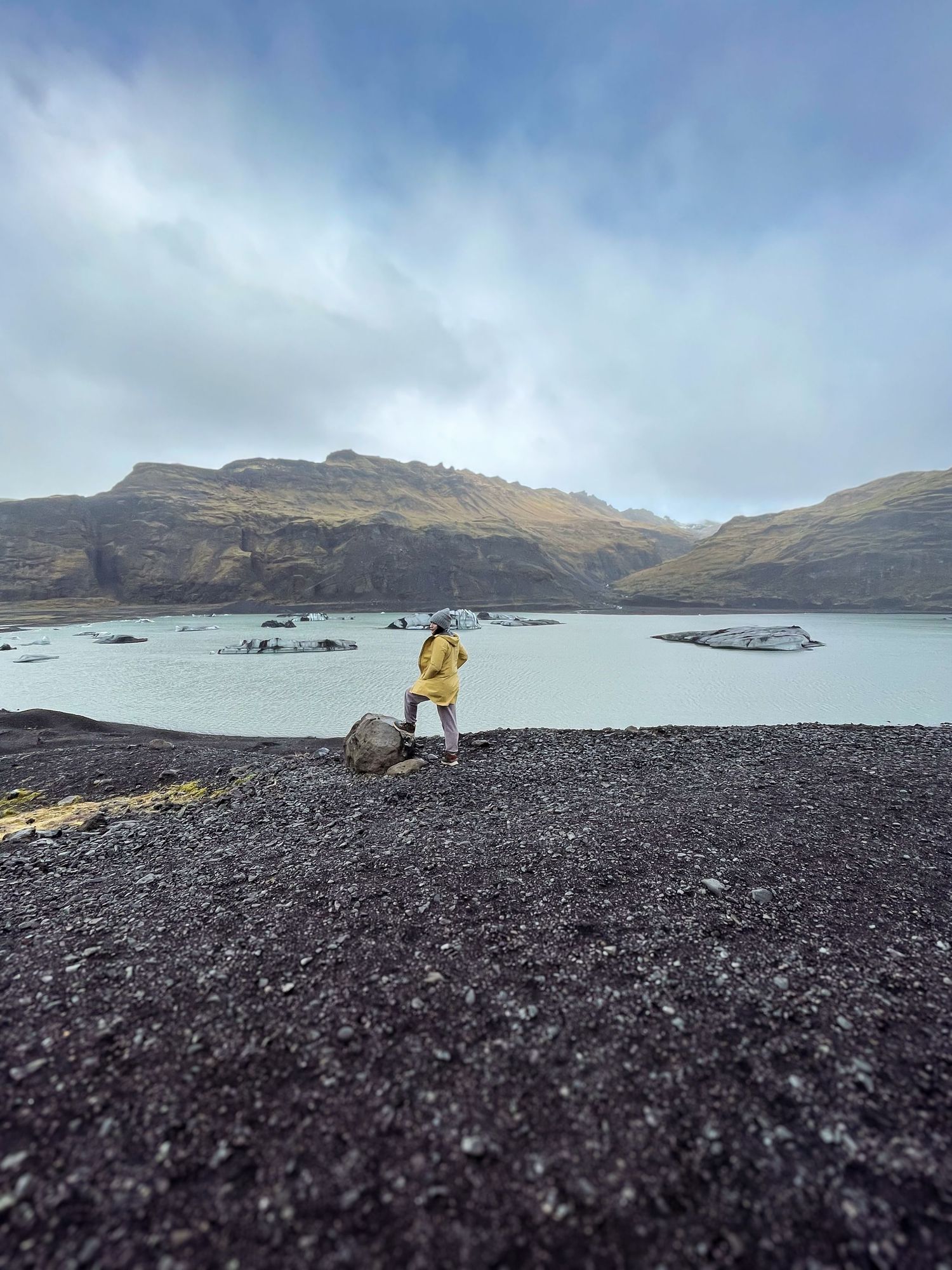 Person in yellow raincoat stands on black volcanic terrain overlooking a glacial lake in Iceland.