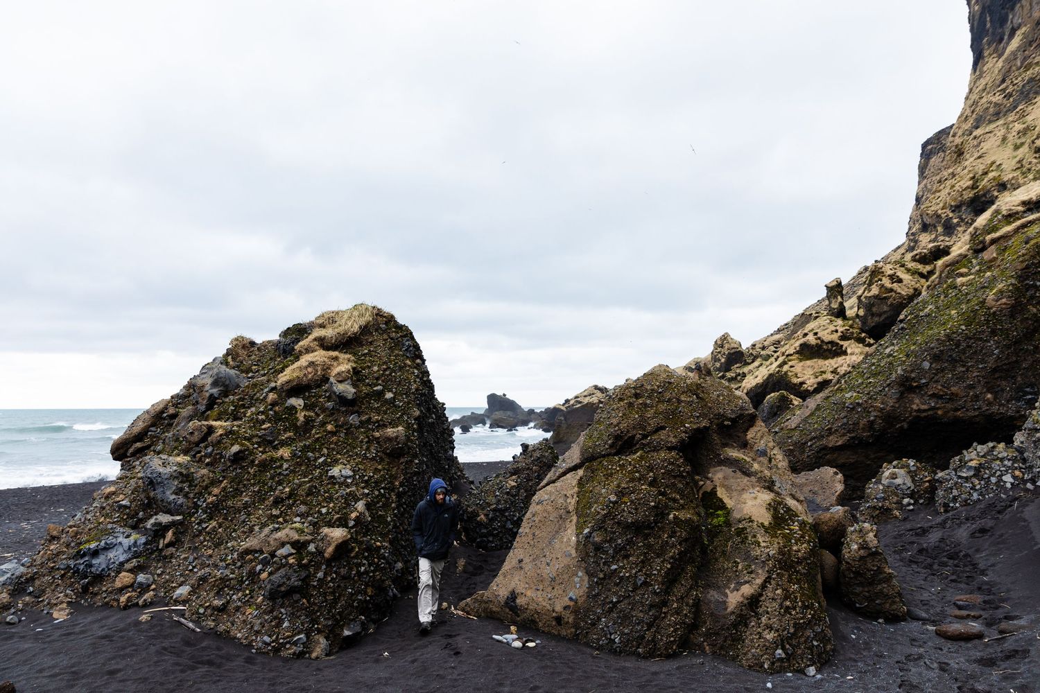 Large rocks and boulders on black sand beach with distant sea stacks visible through gap.