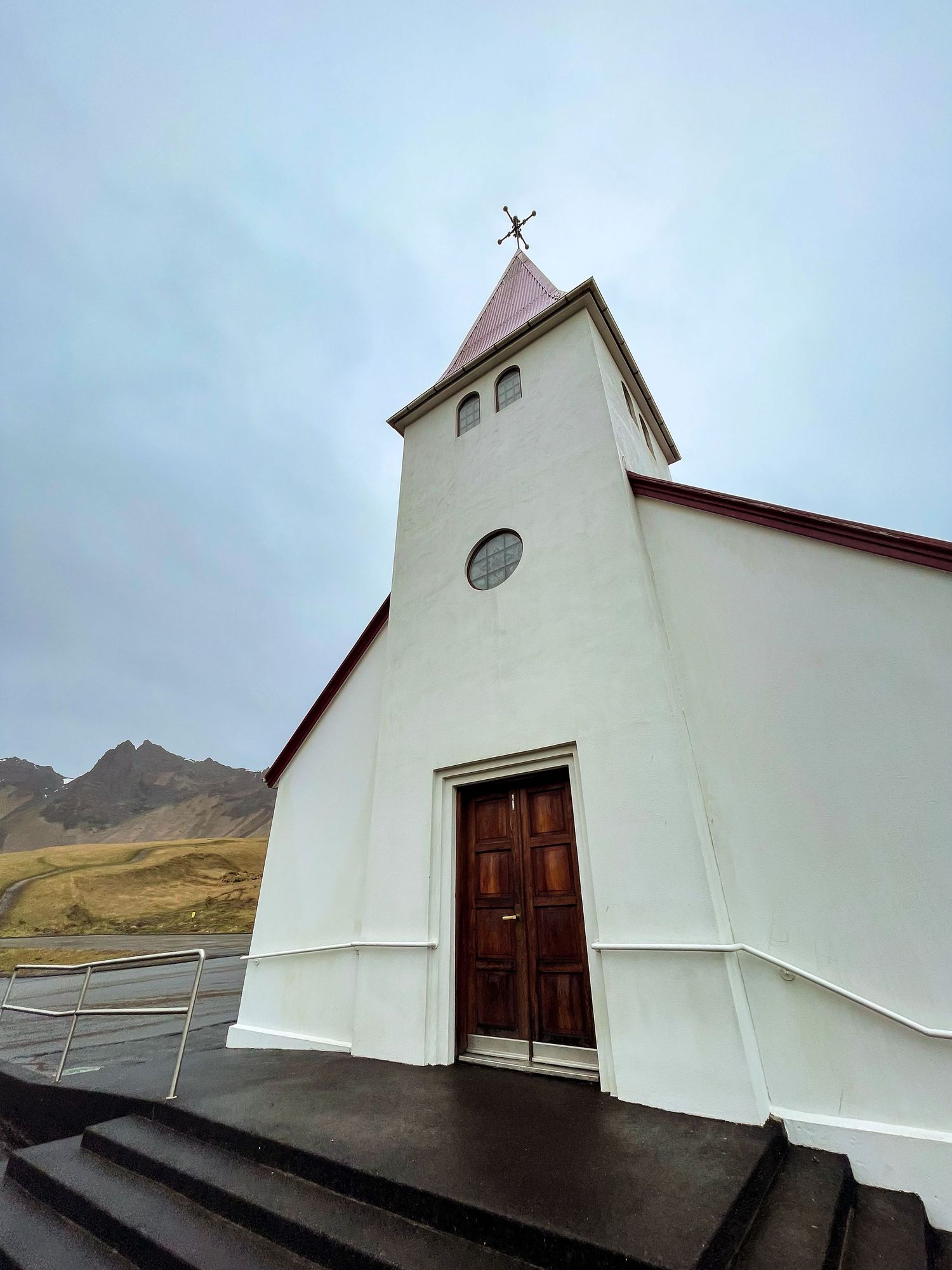 White Nordic church with tall spire and wooden door against mountainous Icelandic landscape.