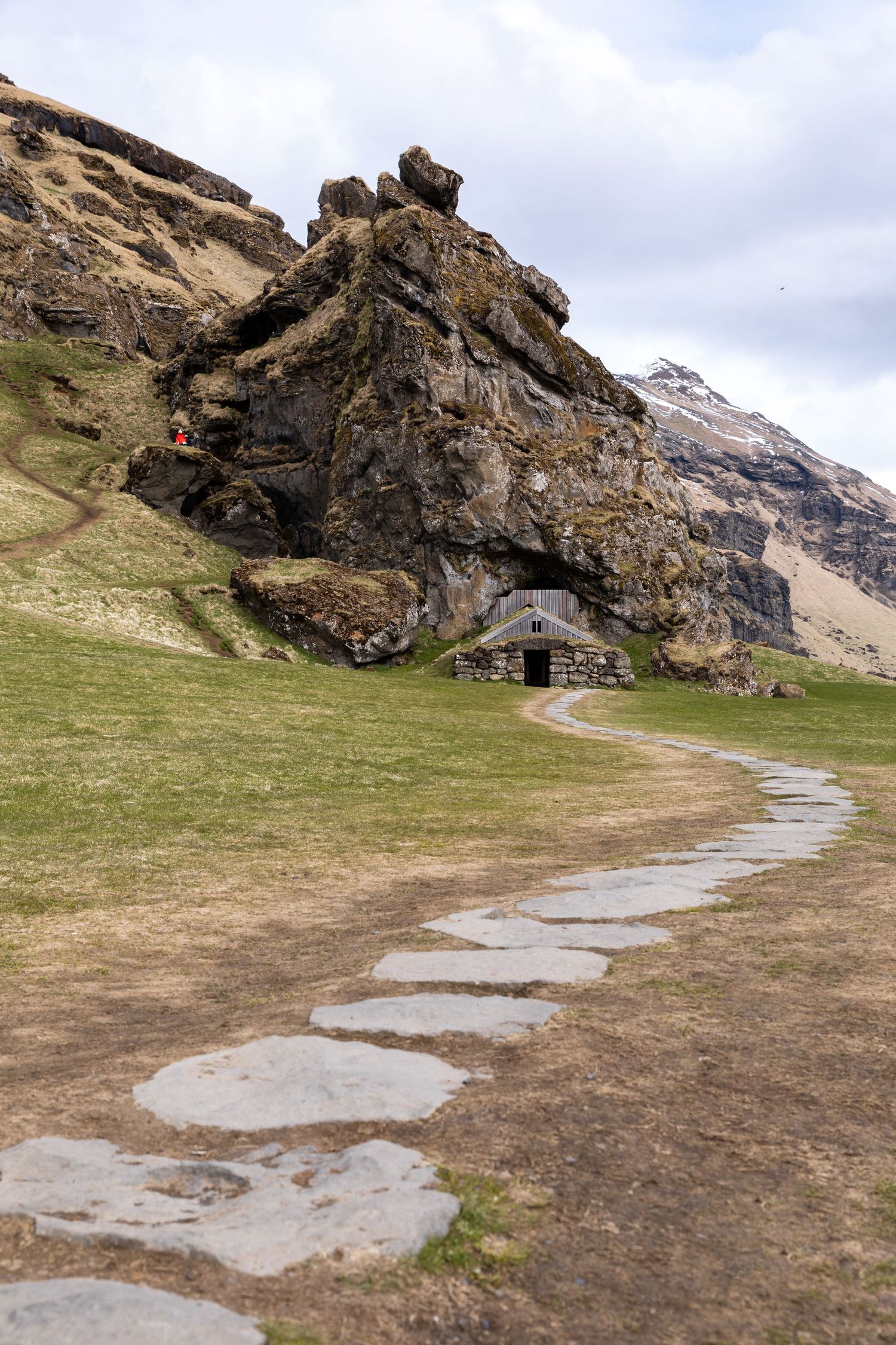 Scenic photo of a troll cave along Route 1 in southern Iceland, with seagulls soaring overhead and a woman in a red coat hiking a mountain t