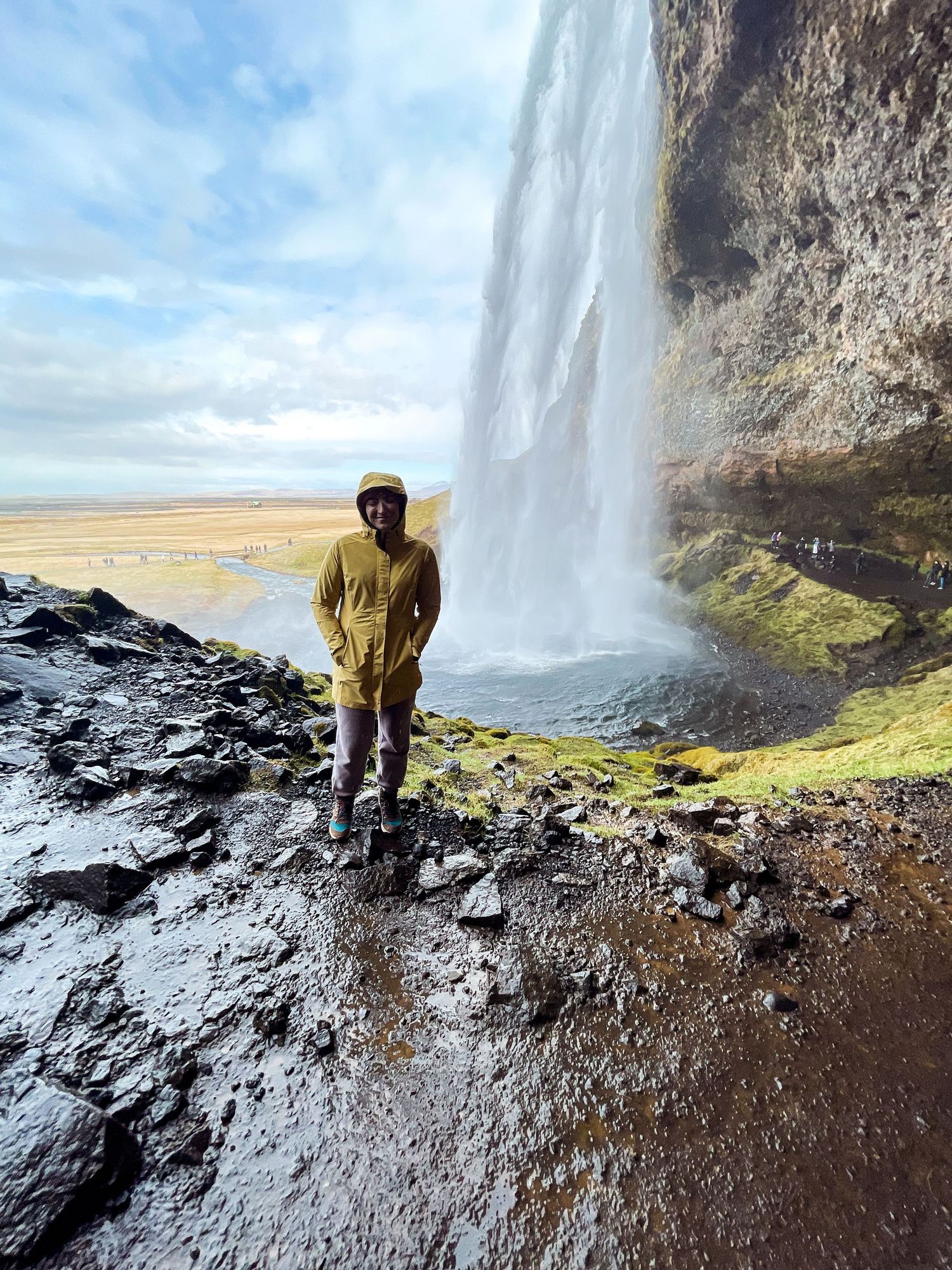 A person in yellow raincoat stands near a massive waterfall cascading down rocky cliffs.