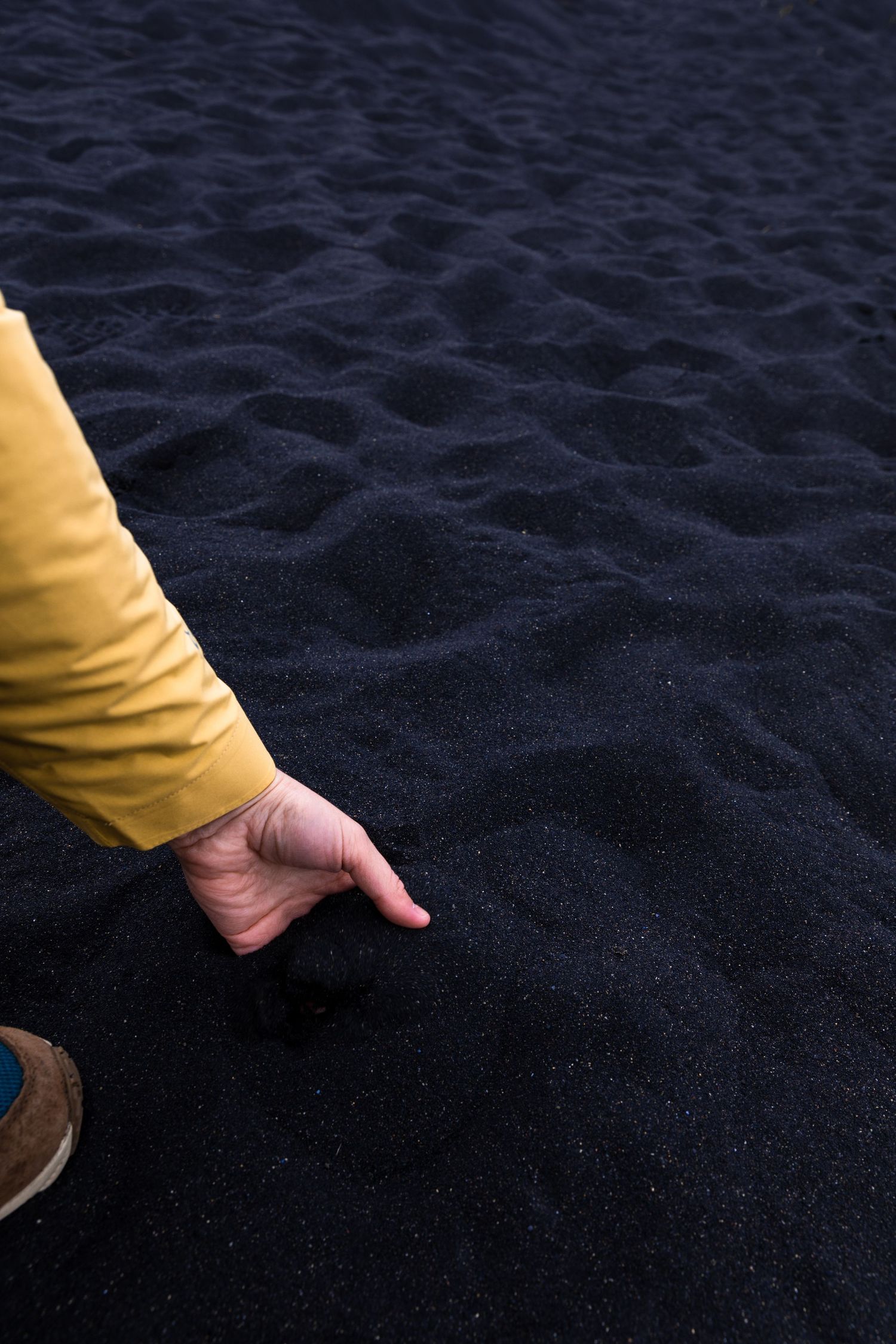 Hand in yellow sleeve touches dark volcanic sand on an Icelandic beach.